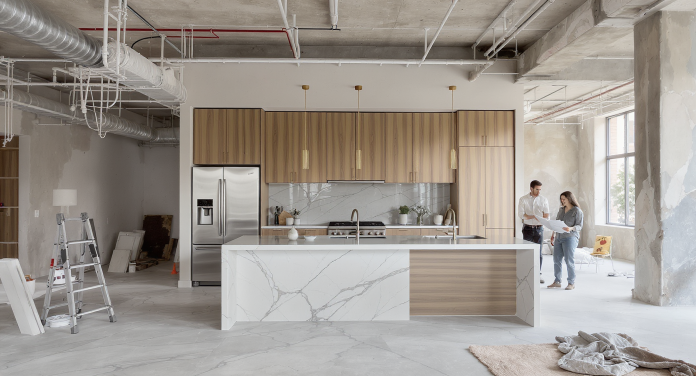 Loft renovation showing intact Henrybuilt kitchen amid open spaces and raw materials, with designer and client reviewing plans.