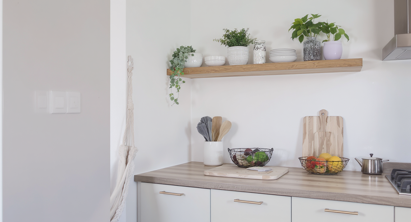 Compact, modern kitchen with floating shelf, fruit hammock holding citrus, and a lower brightly colored net with bananas; countertop holds coffeepot and toaster.