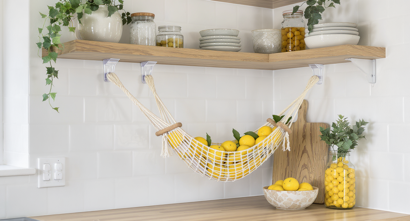 Close-up of a small blue cotton hammock holding lemons and limes, hung in a modern kitchen using removable adhesive hooks under shelves.