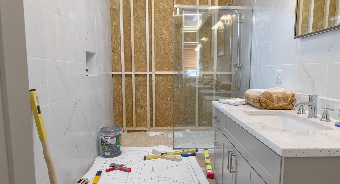 Modern bathroom under renovation featuring an incomplete tiled shower, tools, supplies, and renovation plans on the vanity, illuminated by natural light.