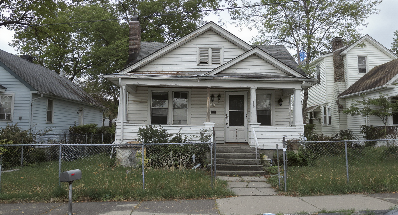 Curbside view of an aged, modest house with peeling paint, worn porch, and patchy yard in a quiet, urban neighborhood, showing renovation needs.
