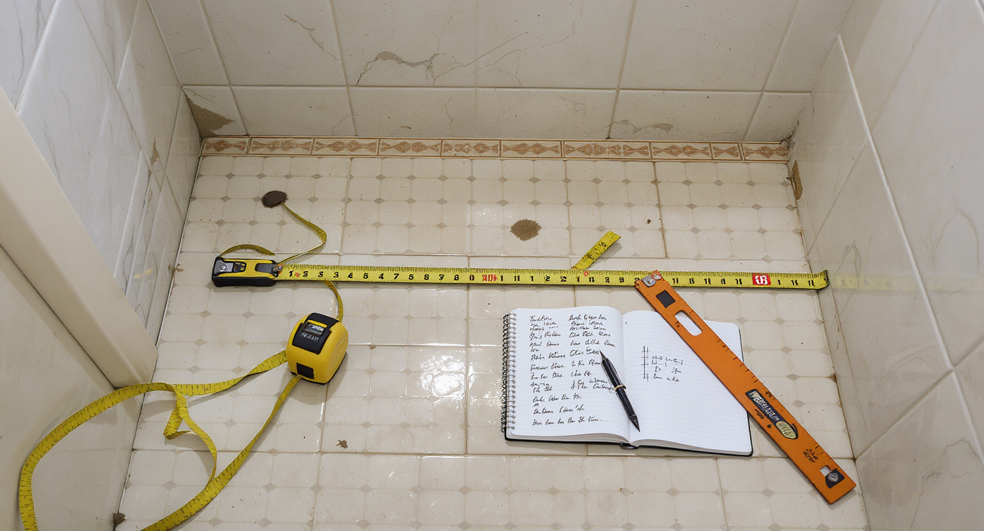 Compact old bathroom with measuring tape across floor, carpenter's square, sketched notebook, and accent tiles stacked, illustrating precise measuring process.