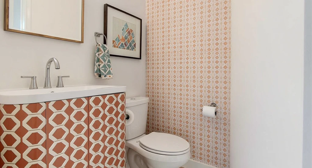 Modern rental bathroom with reddish orange geometric patterned vanity, matching peel and stick wallpaper on feature wall, and accented towels—showing best colors for small bathrooms 2025 and how to combine wallpaper and paint for a rental-friendly makeover.