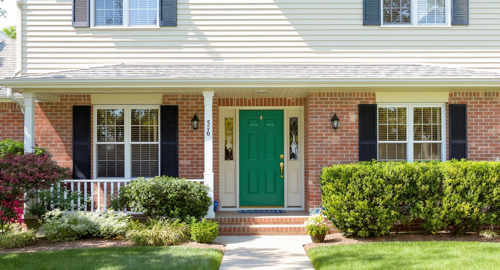 1970s home with unpainted red brick, sand-toned siding, warm green front door, modern fixtures, and lush landscaping. Shows should you paint exterior red brick house alternatives, modern farmhouse curb appeal ideas, and how to choose front door colors.