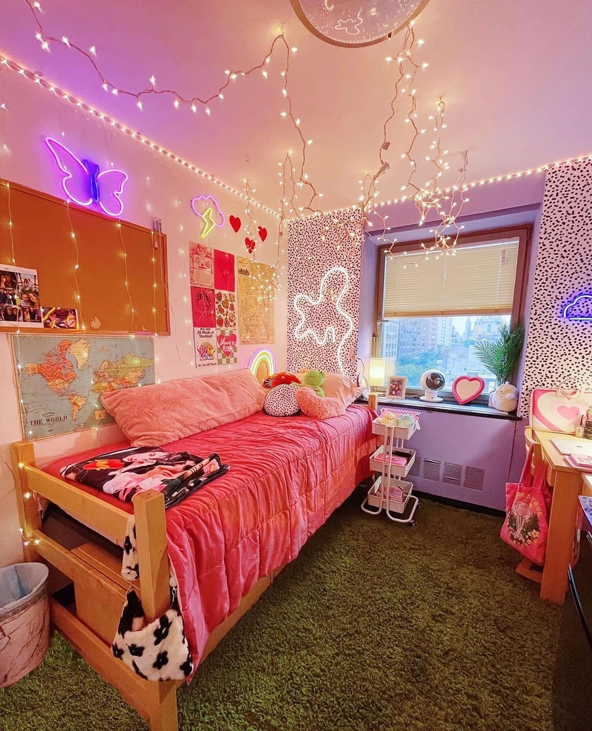 Colorful Y2K-inspired dorm room with pink bedding, fairy lights, neon wall decor, photo collages, plush cushions, a rolling cart, and a bright city view.