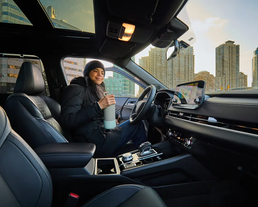 Woman in winter coat and hat holding a water bottle inside a modern car with city buildings outside.