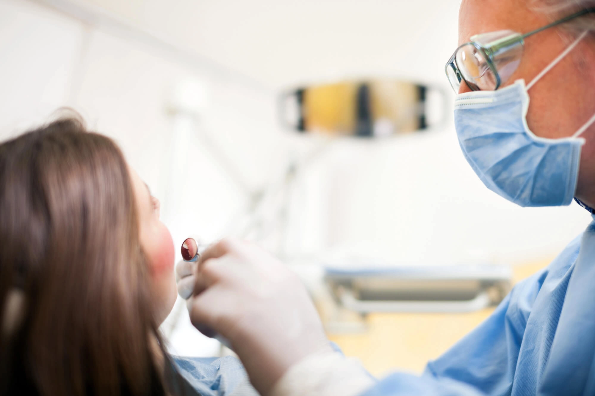 Dentist examining a patient's mouth using a dental mirror and wearing blue scrubs and gloves in a clinical setting