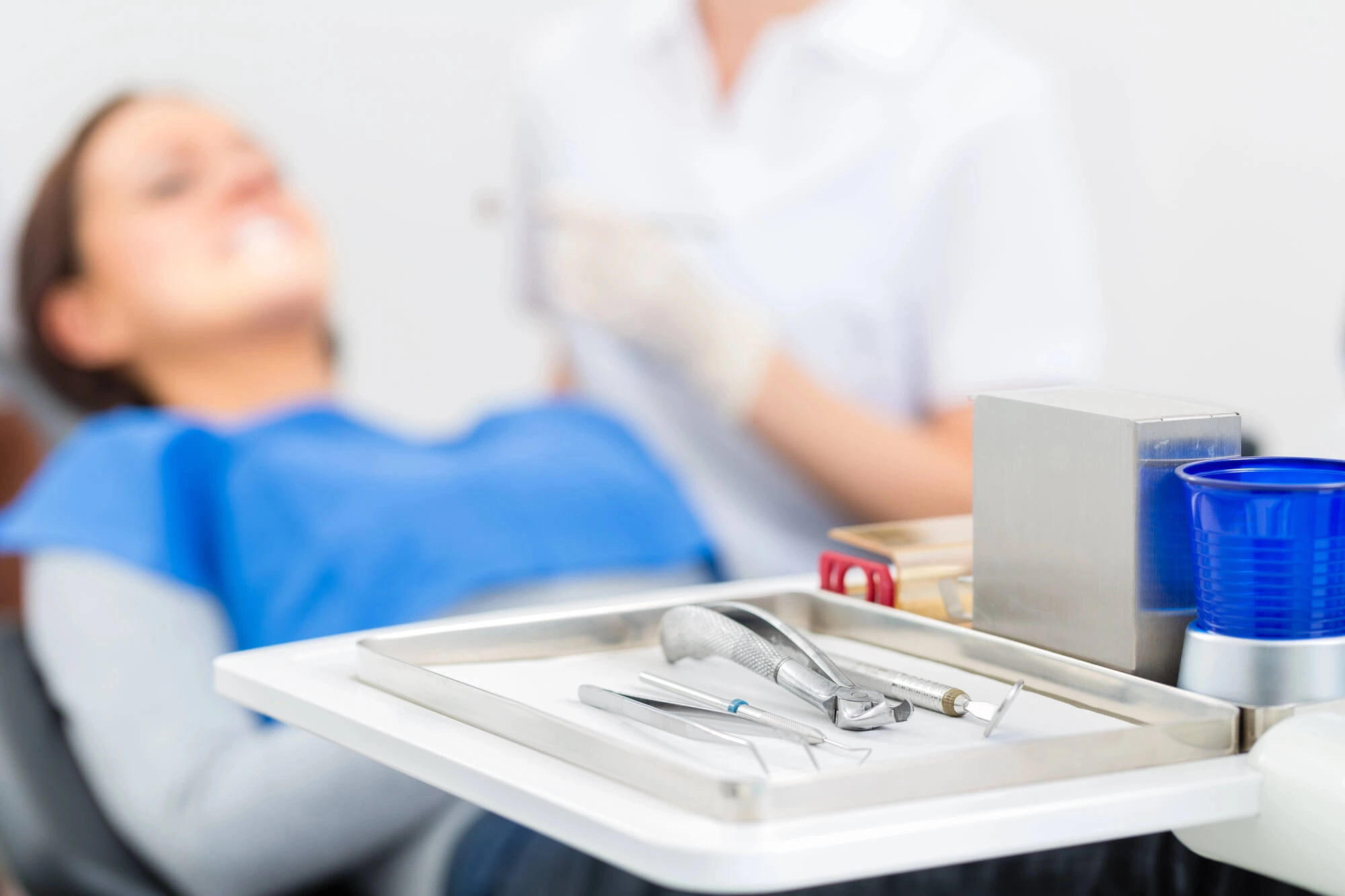 Dental tools on a tray in the foreground with a patient lying back and a dentist in the background, preparing for a dental procedure