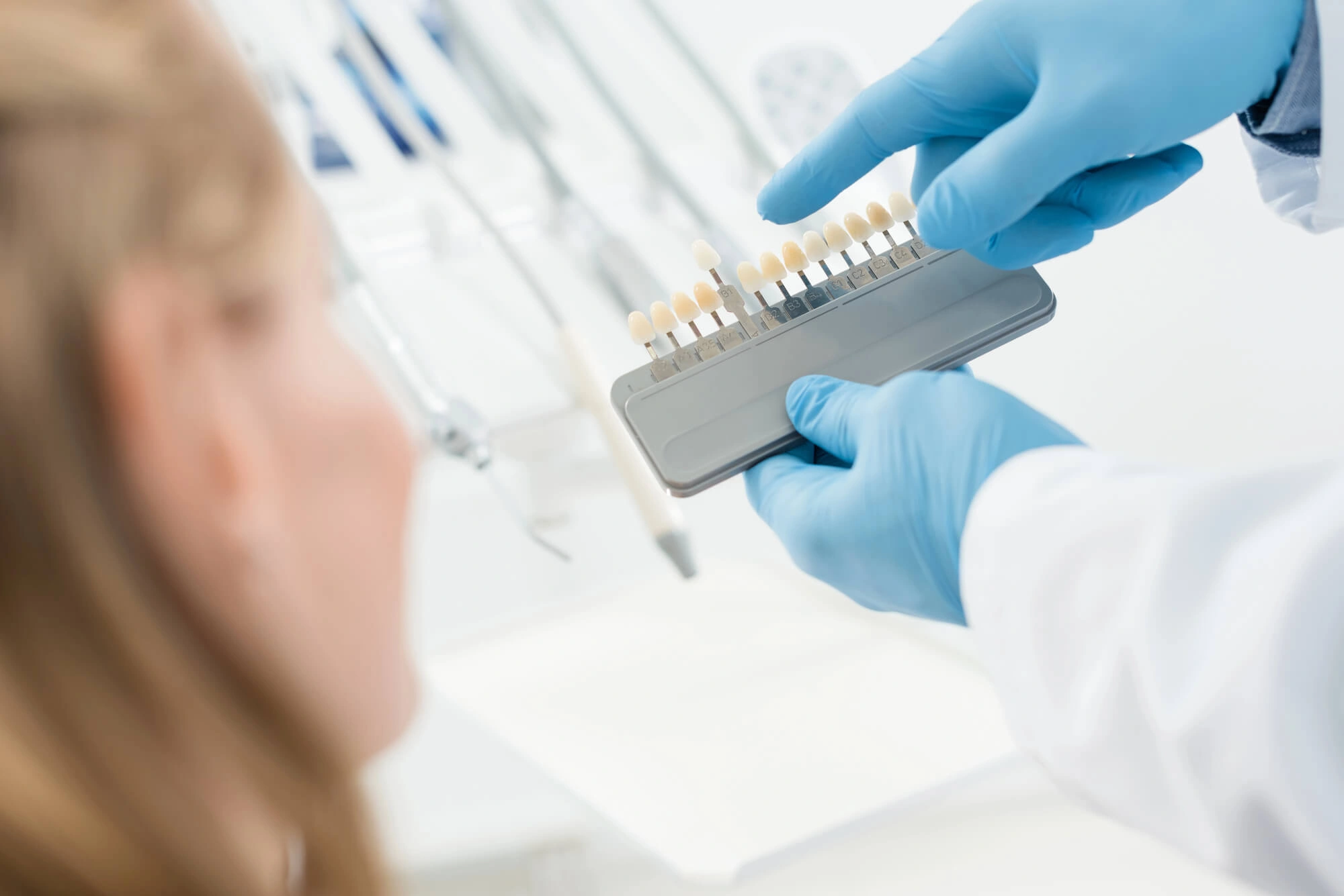 A dentist in blue gloves holds a tooth color shade guide, showing it to a patient in a dental clinic. The scene is professional and clinical