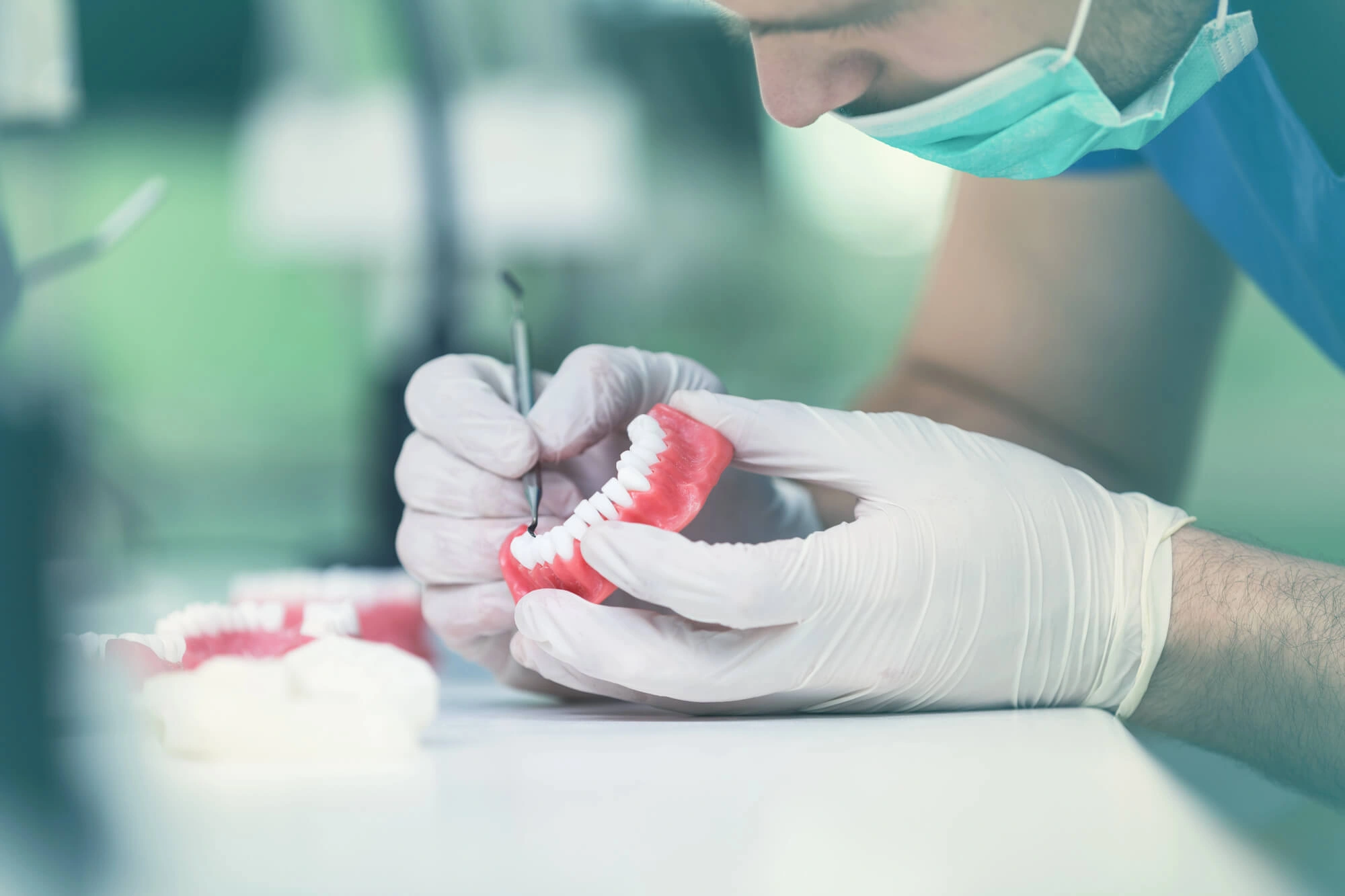 A dental technician wearing gloves and a mask works meticulously on a set of dentures using a precision tool, conveying focus and care