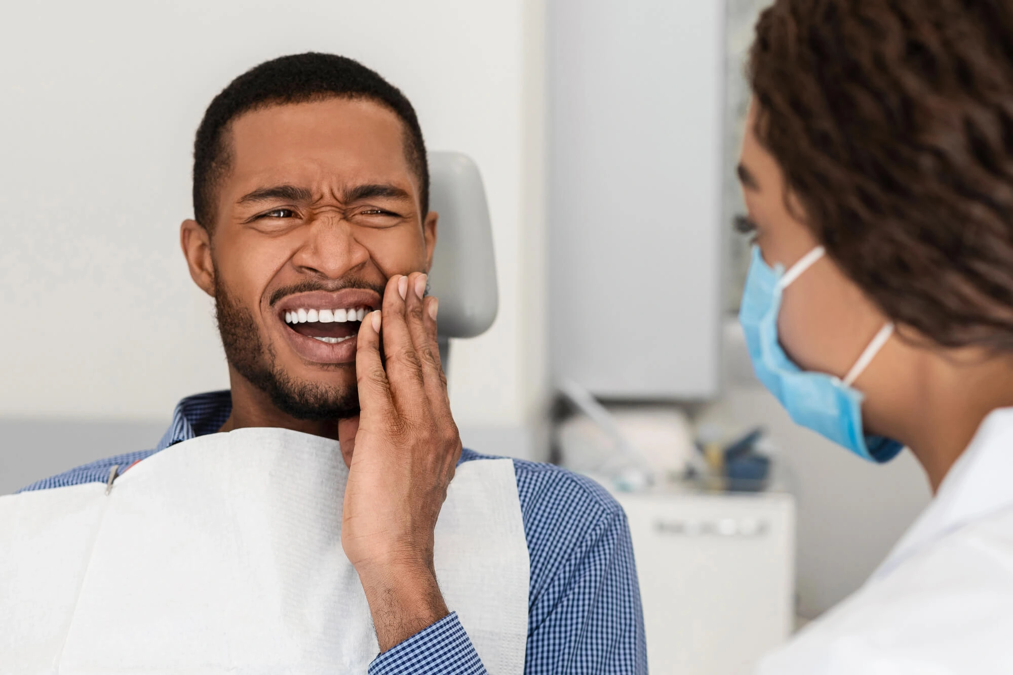 A man in a dental chair winces in pain, holding his cheek, while a dentist, wearing a mask, looks on with concern. The setting is clinical