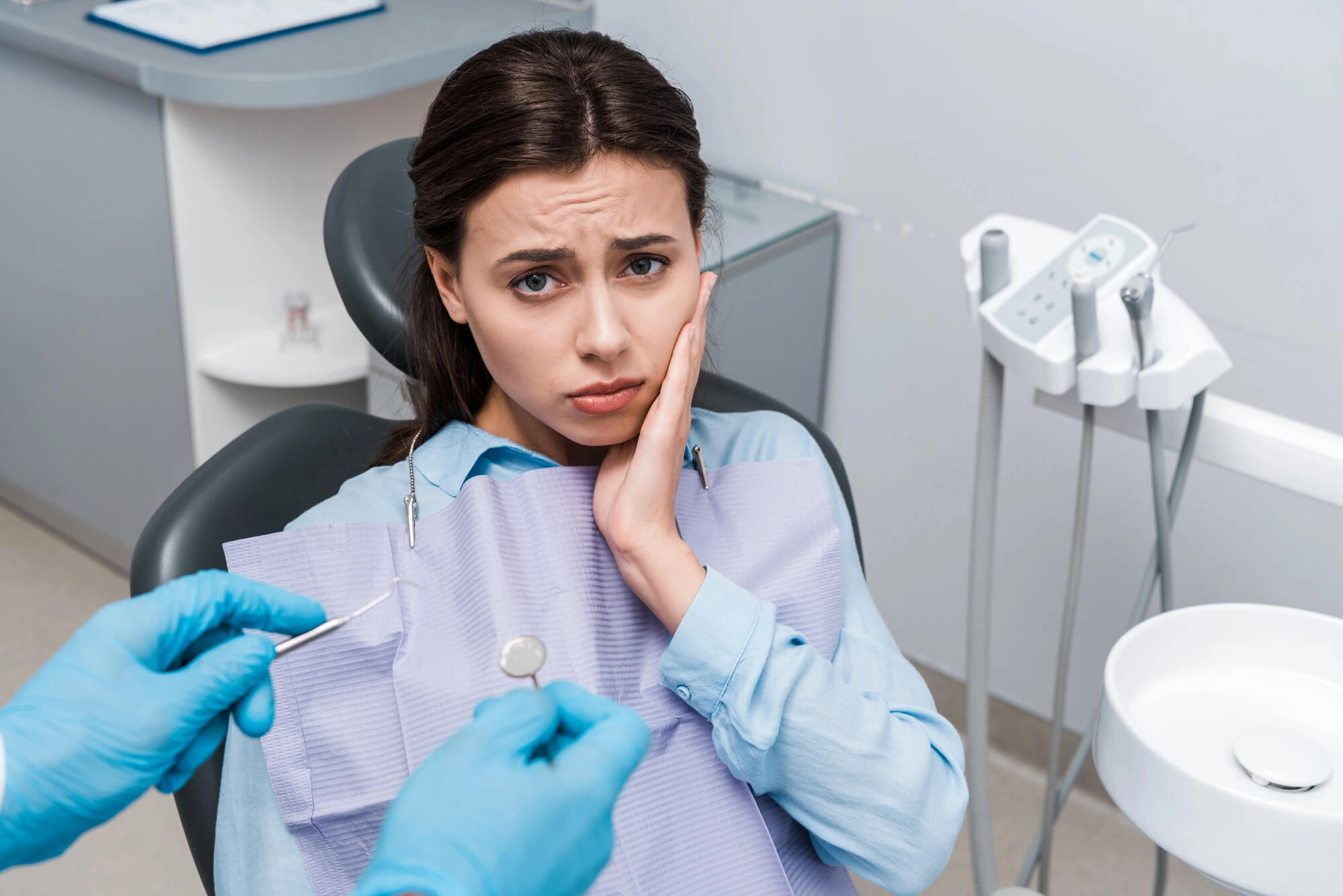A woman looks concerned, holding her cheek in a dental chair. A dentist in gloves holds tools nearby. The setting is a bright dental office