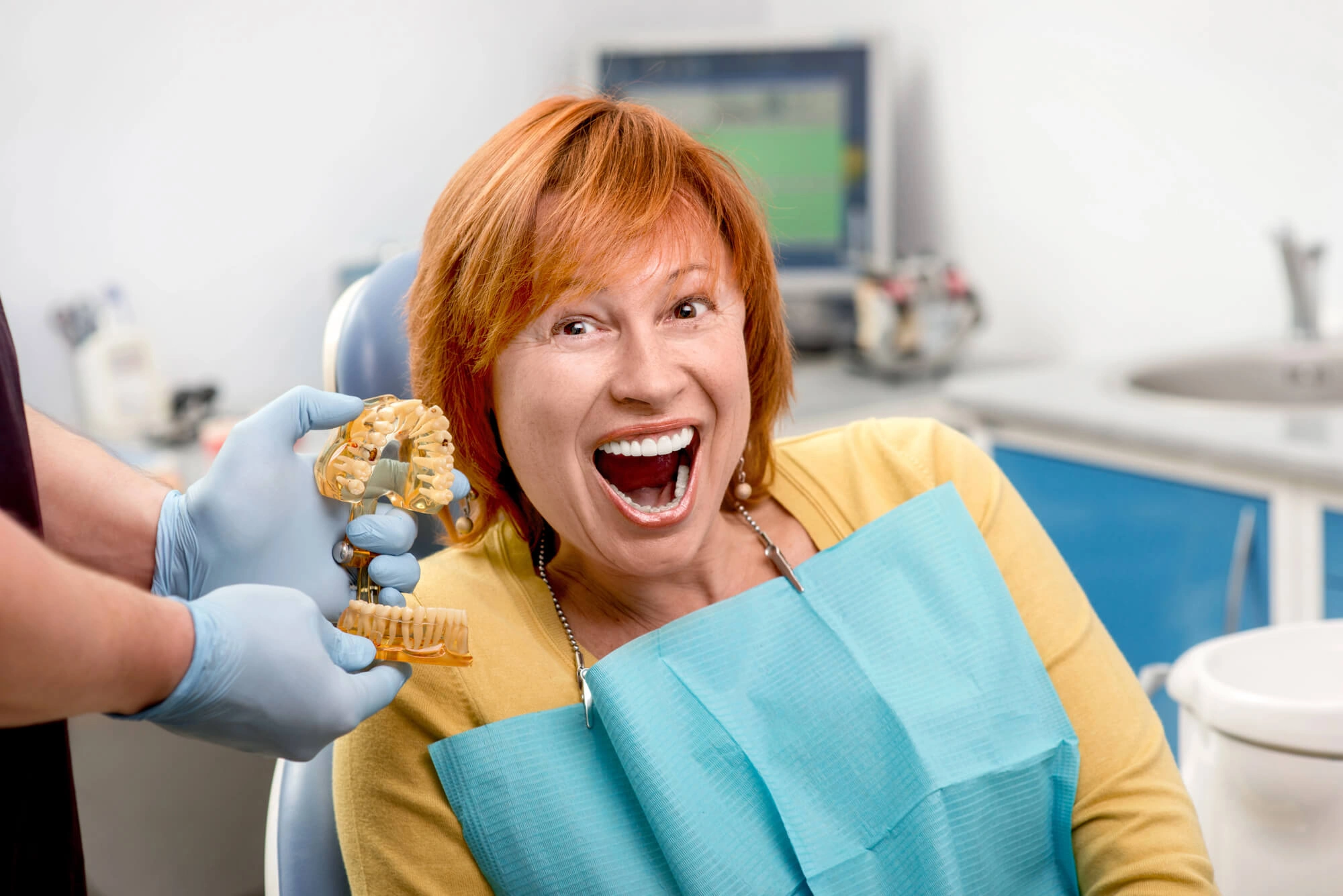 Dentist wearing gloves shows a dental model to a patient sitting in the dental chair with a blue bib in a clinic