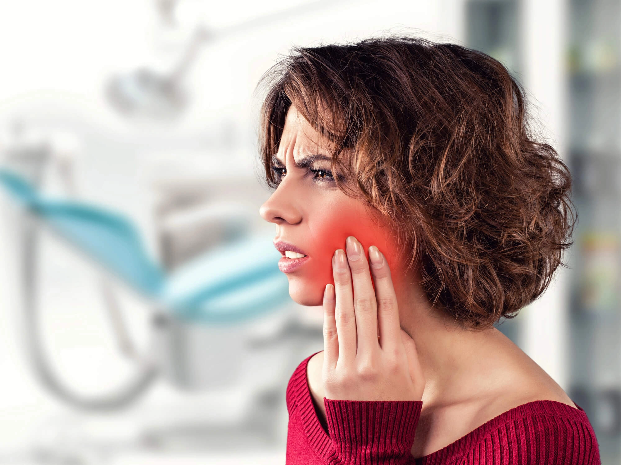 A woman in a dental office holds her jaw with a pained expression. Her cheek is visibly red, indicating discomfort or a toothache. Dentist chair blurred in background