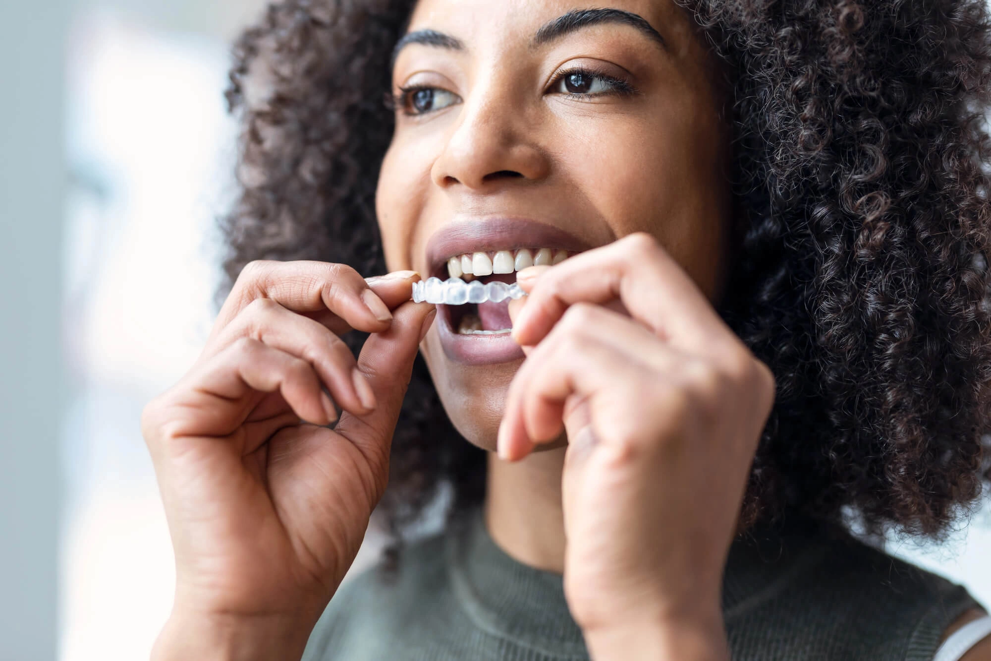 A person with curly hair is smiling while inserting a clear dental aligner over their teeth. The scene conveys a sense of care and confidence