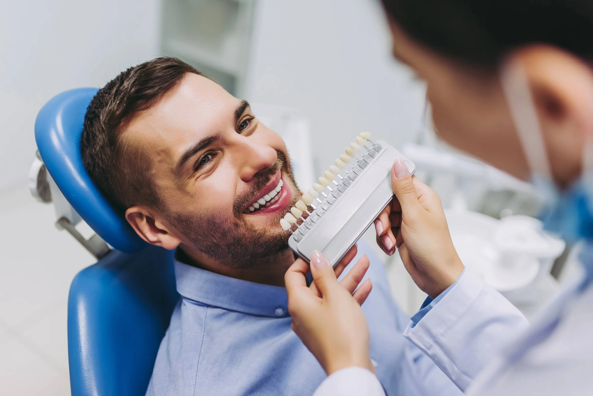 A man in a dental chair smiles as a dentist holds up a tooth color chart to his teeth. The setting is clinical, conveying professionalism and care