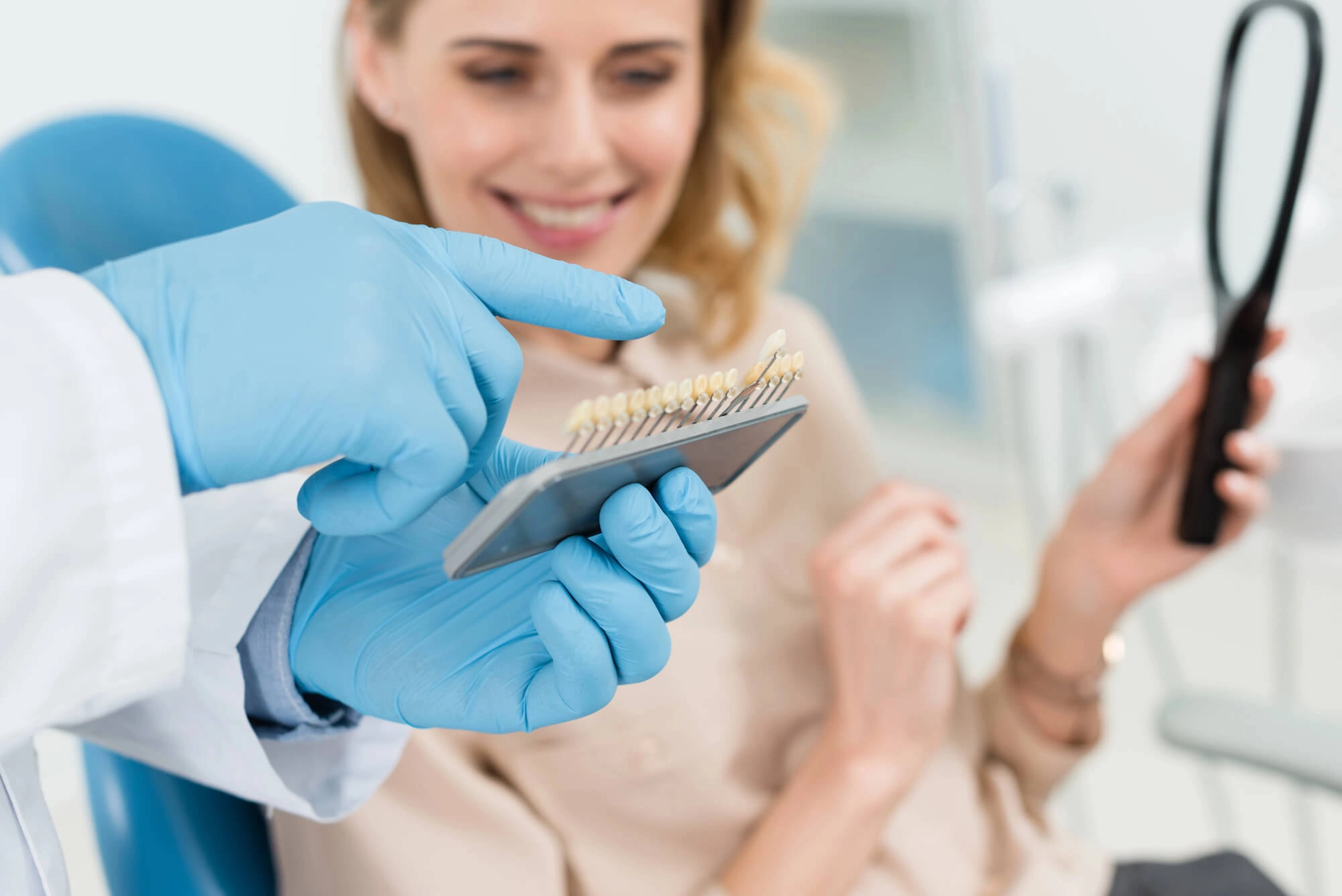Dentist with blue gloves holds a shade guide, showing tooth colors to a smiling woman in a dental chair holding a mirror, ensuring a relaxed tone