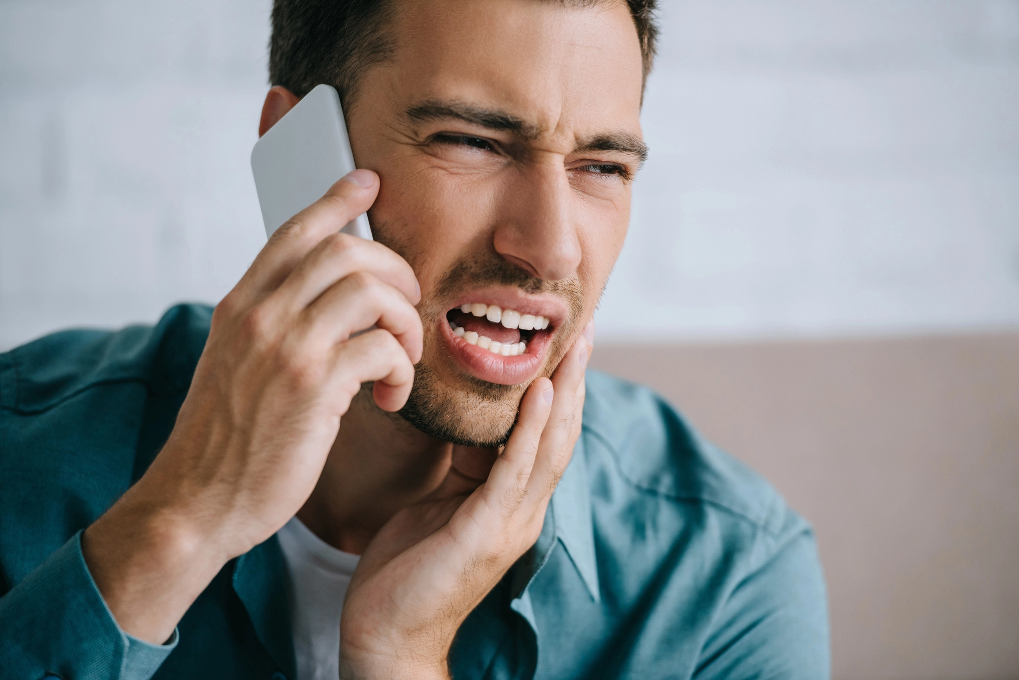 A man holding his jaw in pain talks on the phone, wearing a blue shirt against a neutral background. The expression shows discomfort and urgency