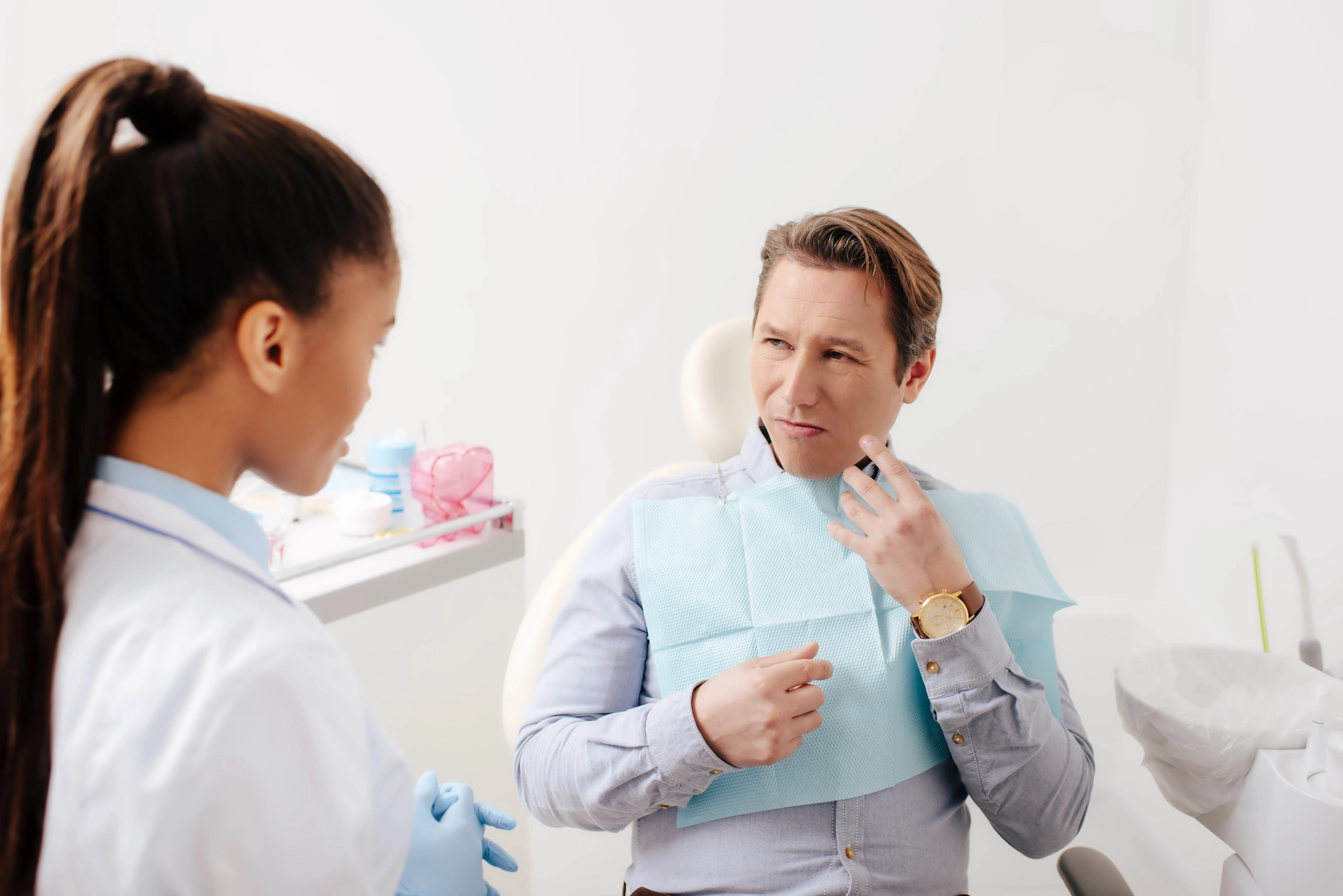 Man sits in a dental chair, touching his jaw, looking concerned. Dentist stands nearby, listening and ready to assist in a clean, bright office