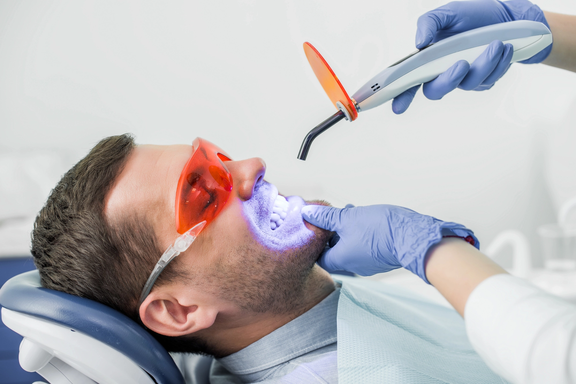 A dentist in gloves uses a curing light on a patient's teeth during a dental procedure. The patient wears protective orange goggles, conveying a clinical setting