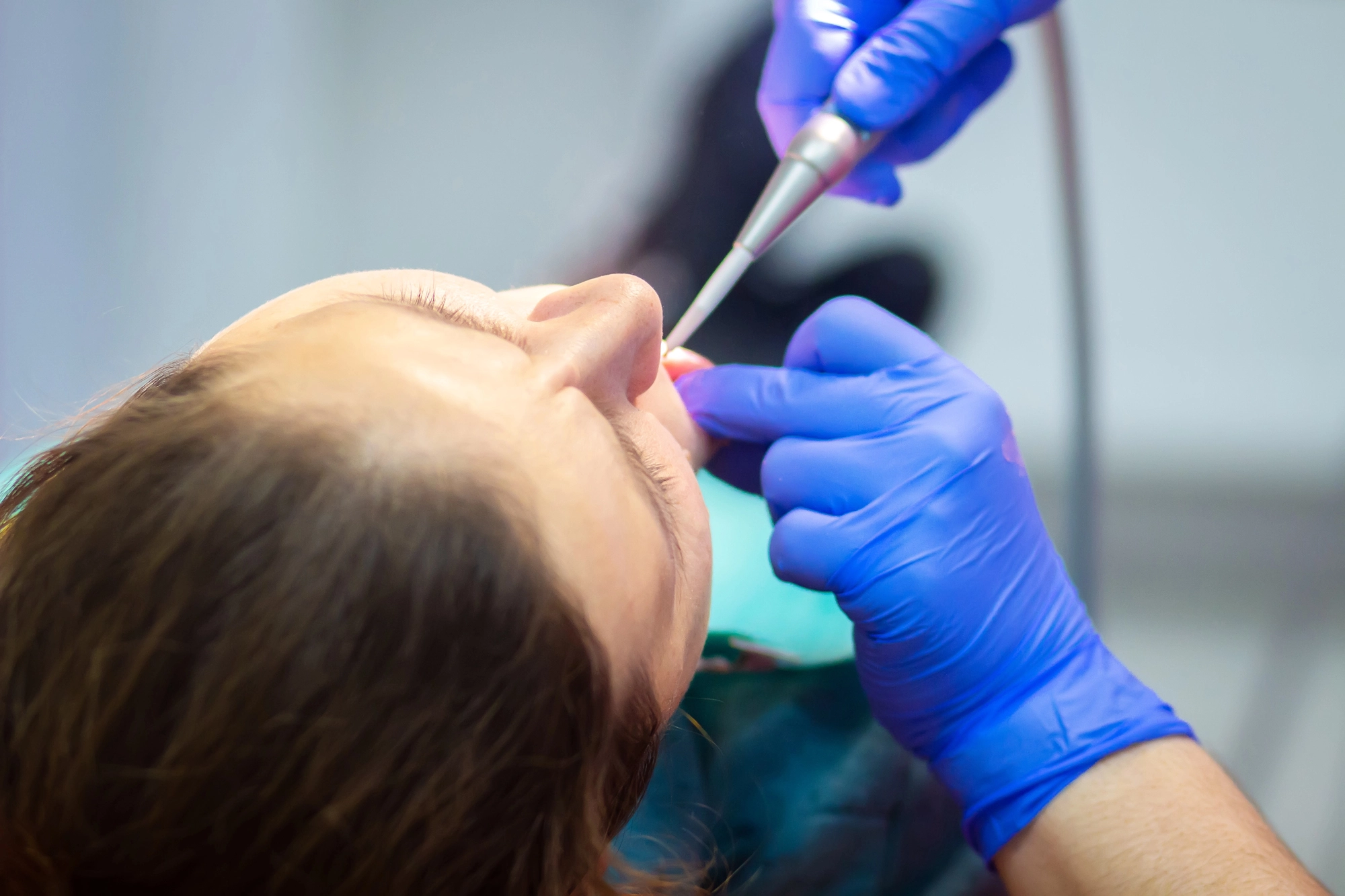 A dentist in blue gloves uses a dental tool to work on a patient's teeth. The patient lies back, looking relaxed, in a clinical setting