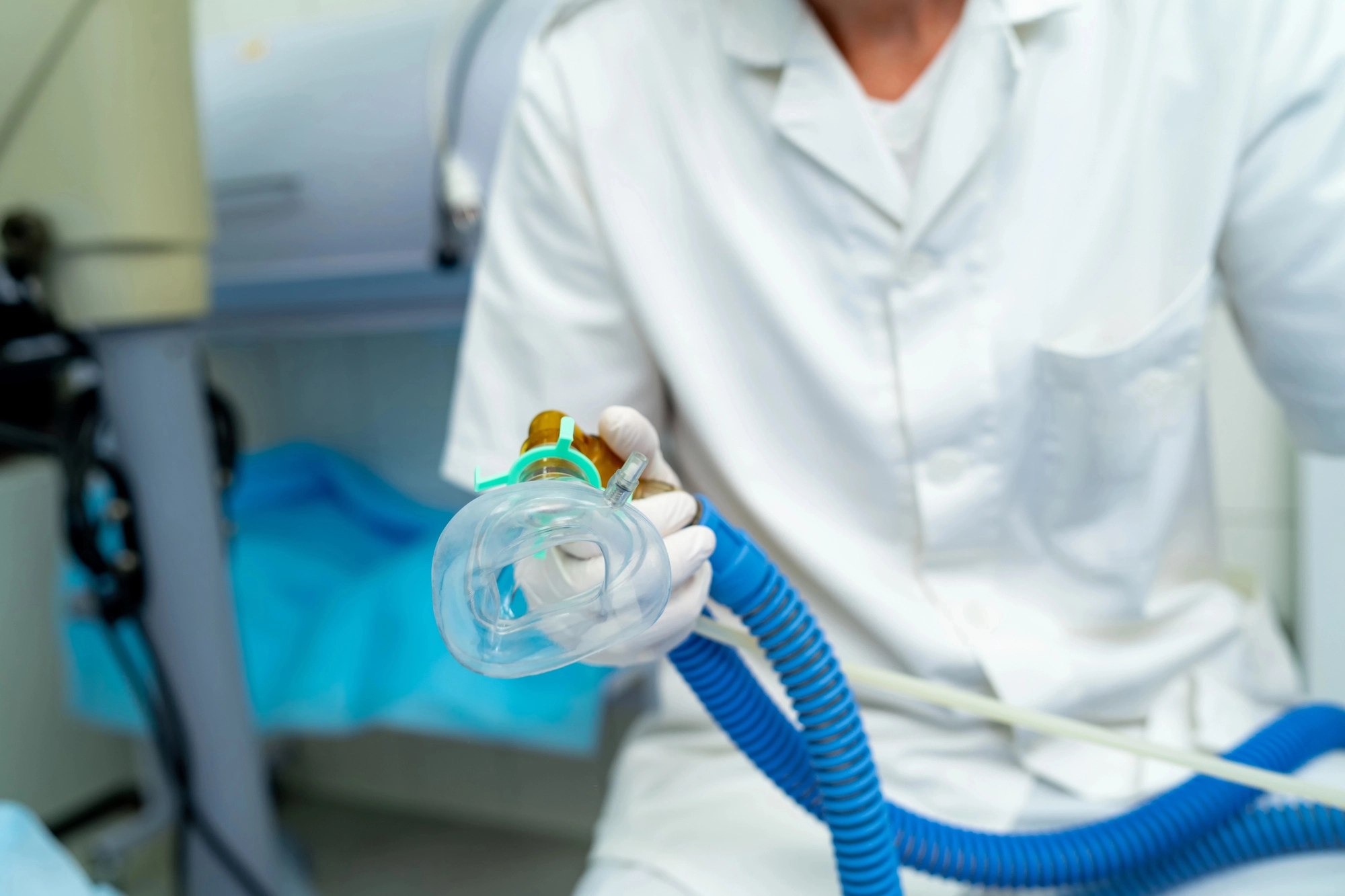 A healthcare professional in white coat and gloves holds a clear ventilation mask with a blue tube in a clinical setting, conveying preparedness and care