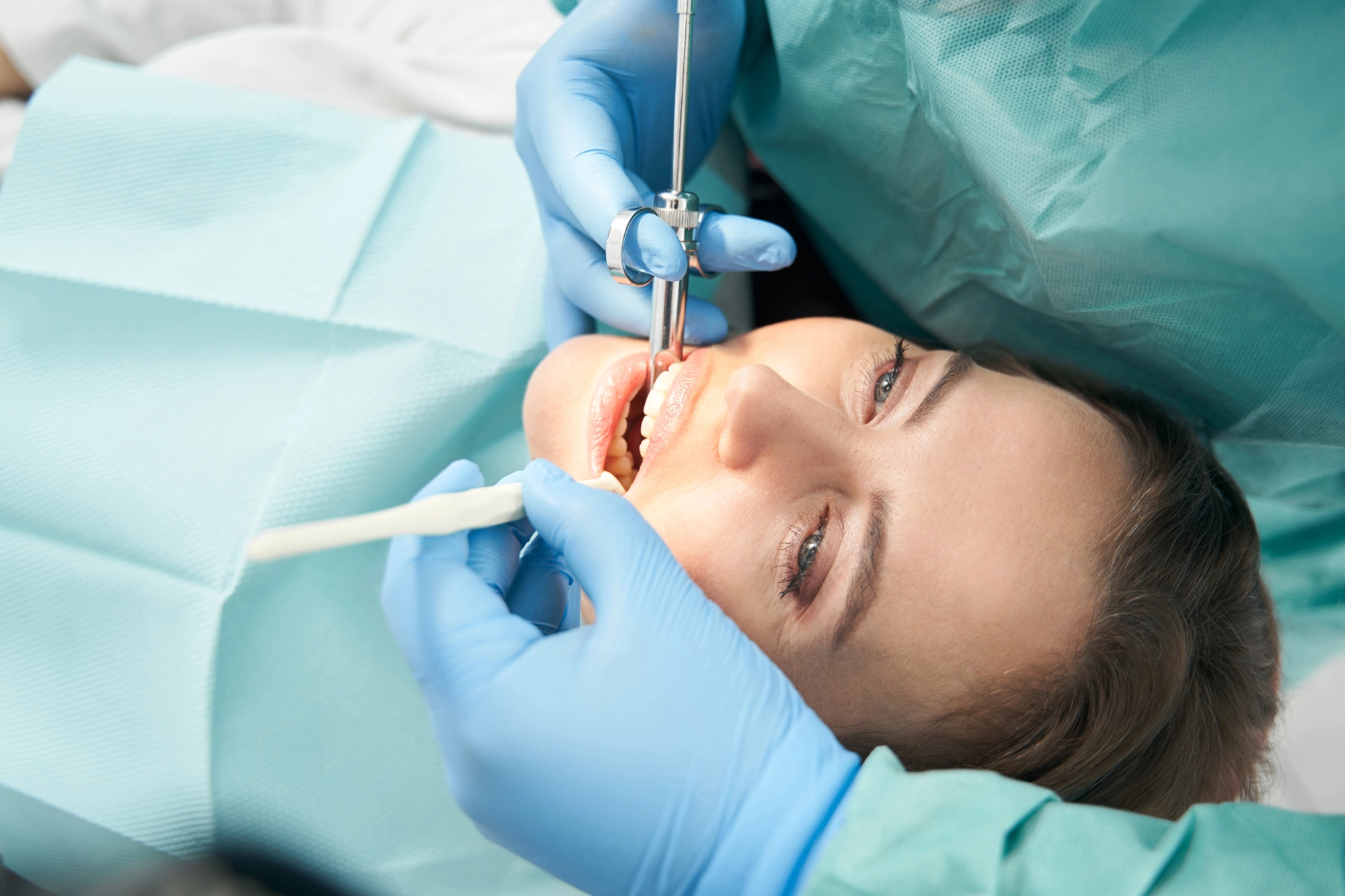 A dentist wearing blue gloves works on a patient's teeth using dental tools. The patient is reclined and covered with a teal protective drape