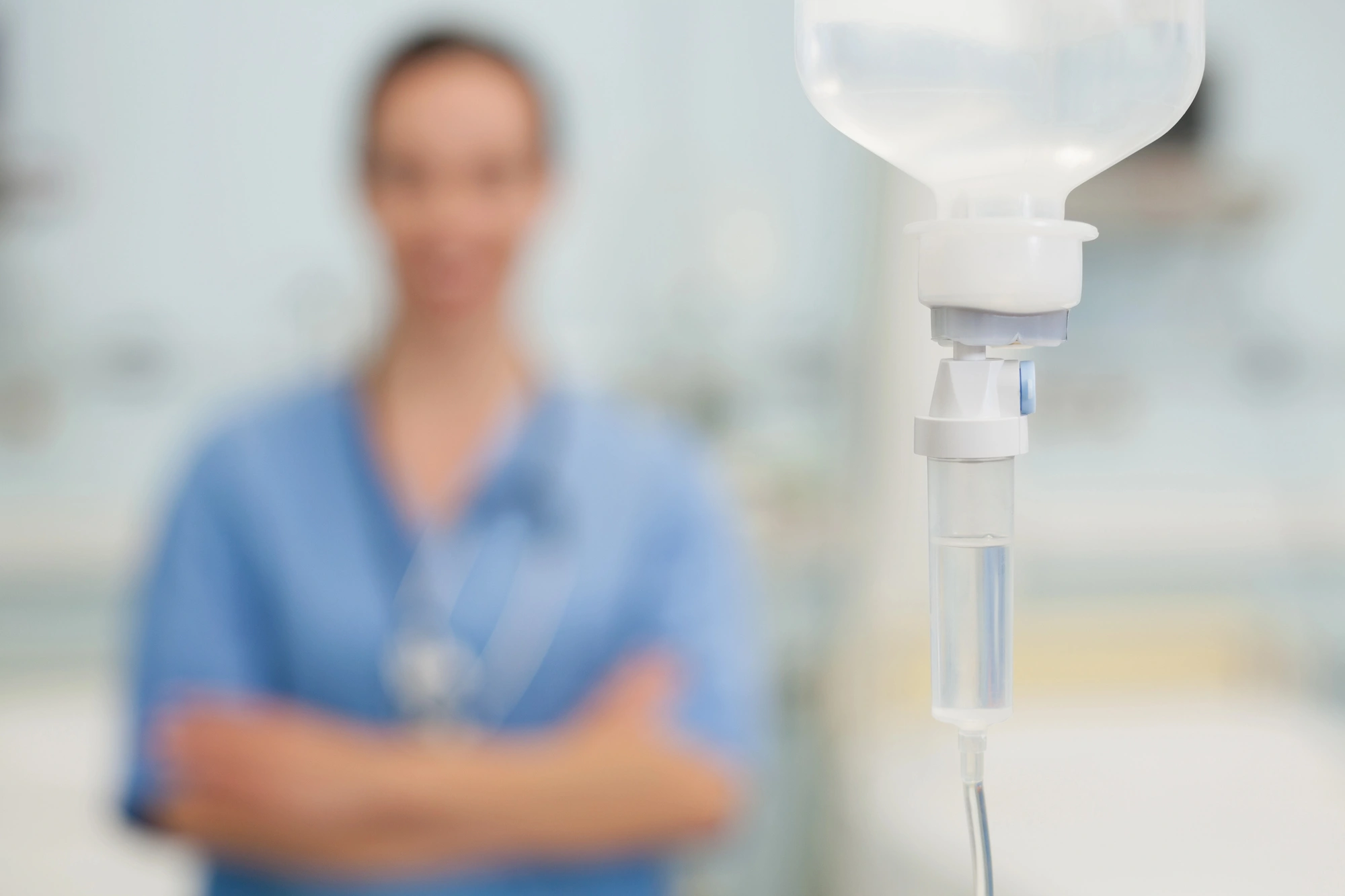 A blurred medical professional stands in the background with crossed arms, while a sharp IV drip hangs in the foreground, conveying a healthcare setting