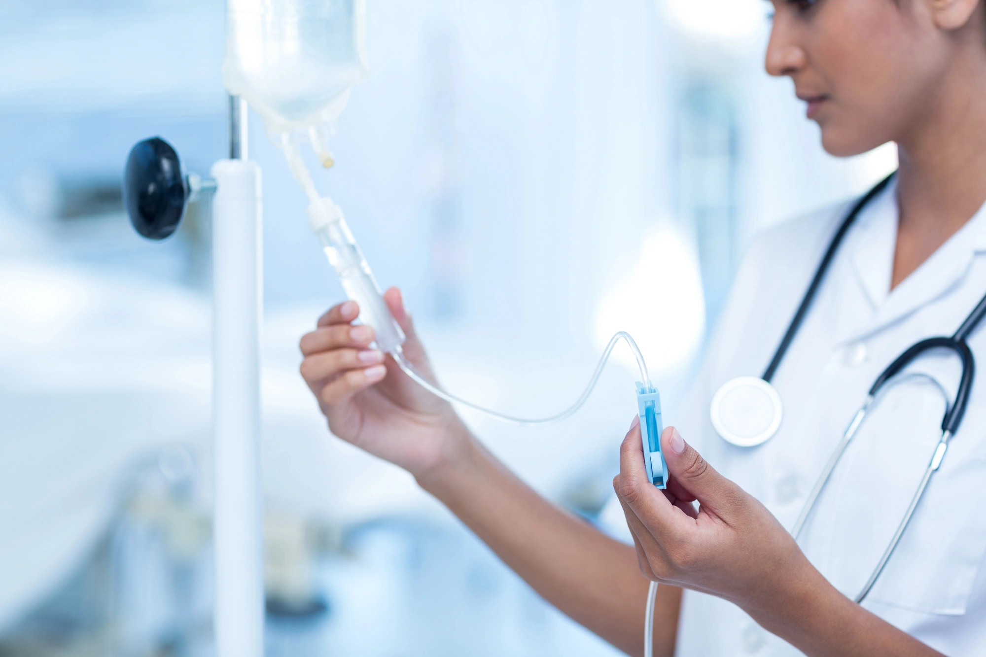 A nurse in a white uniform adjusts an IV drip in a hospital setting. The image conveys a sense of focus and professionalism in a clinical environment