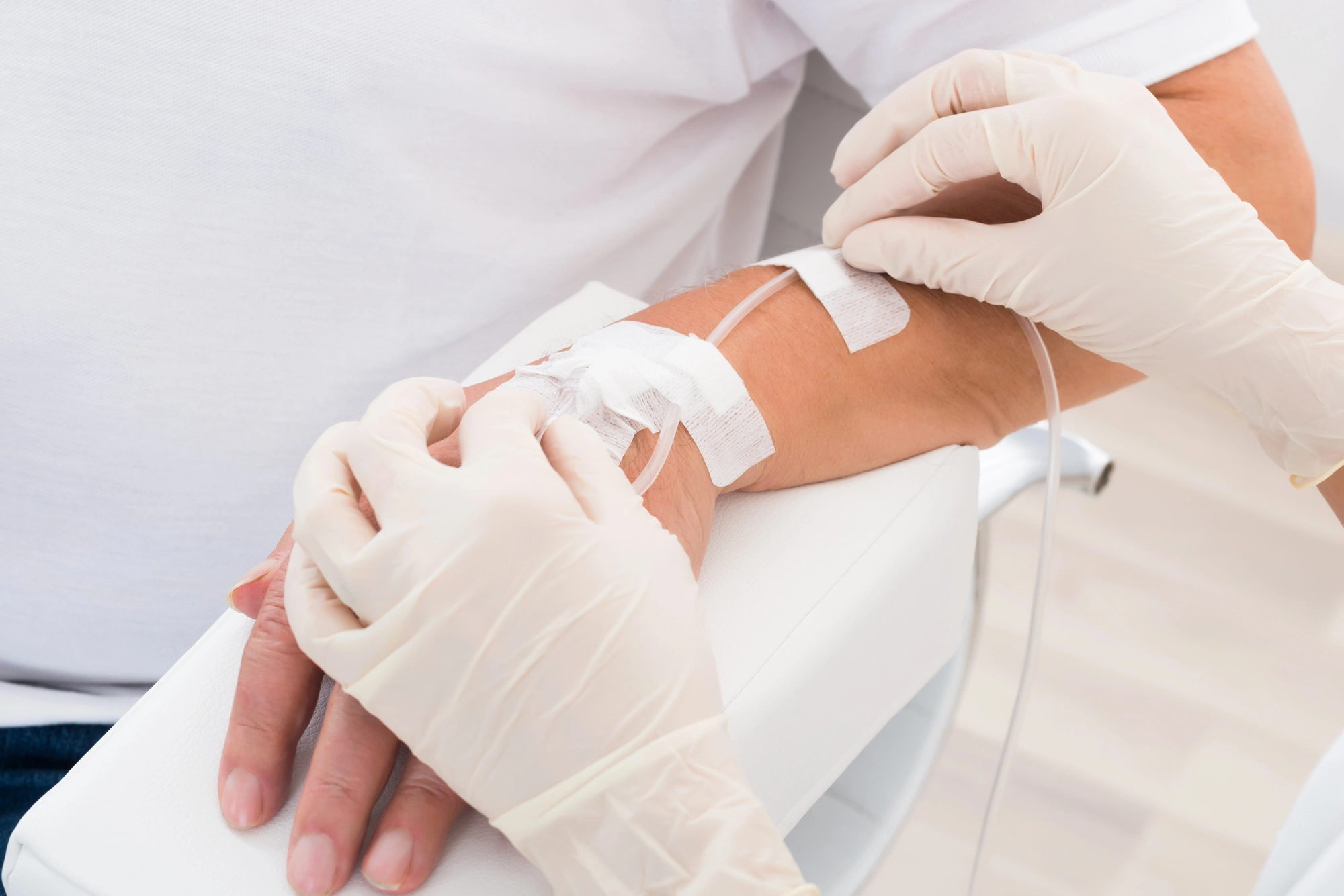Hands in medical gloves carefully insert an intravenous line into a patient's arm, fixed with white tape, on a white chair. The tone is clinical and calm