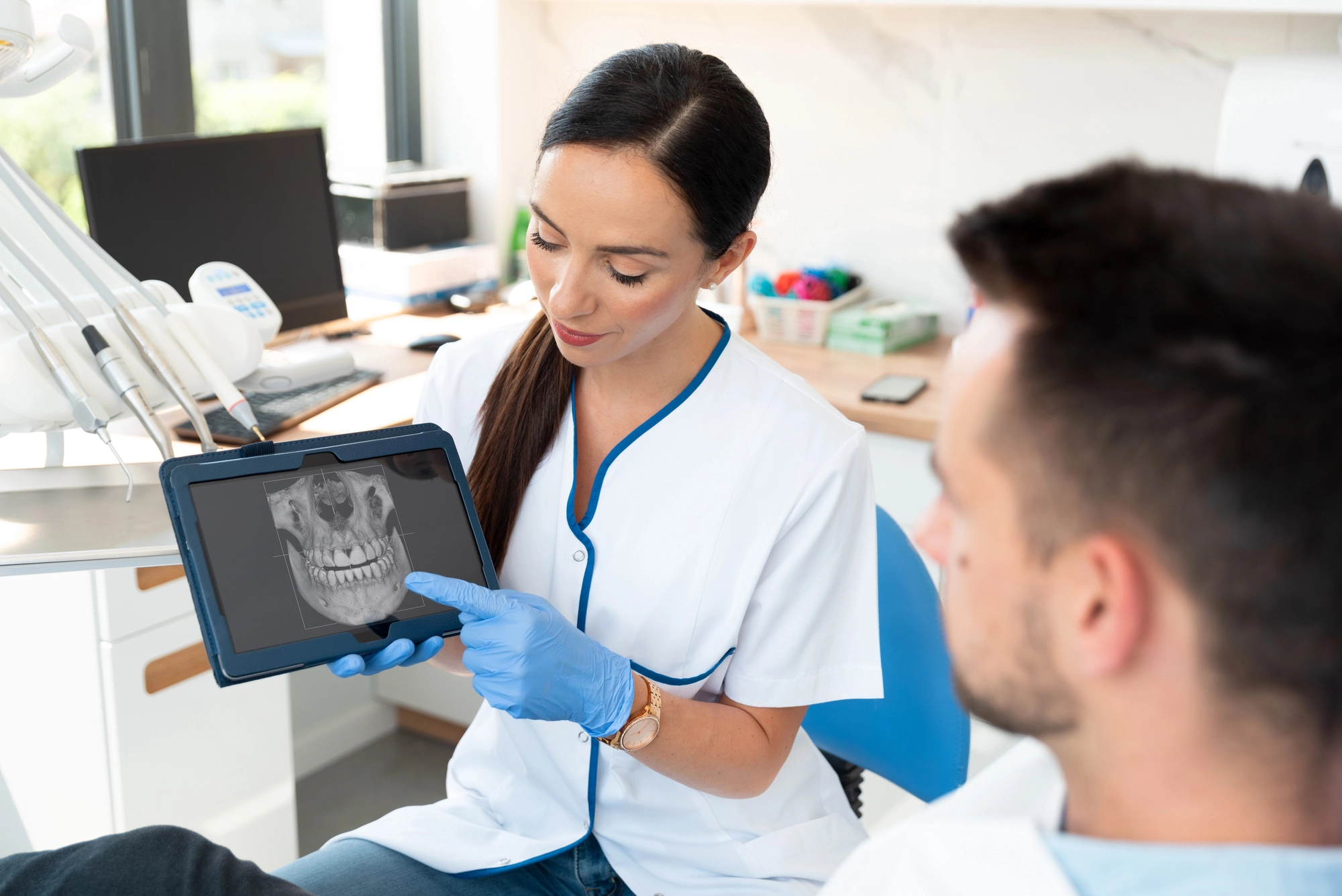 Dentist in white coat and blue gloves showing a dental X-ray on a tablet to a seated patient in a clinic