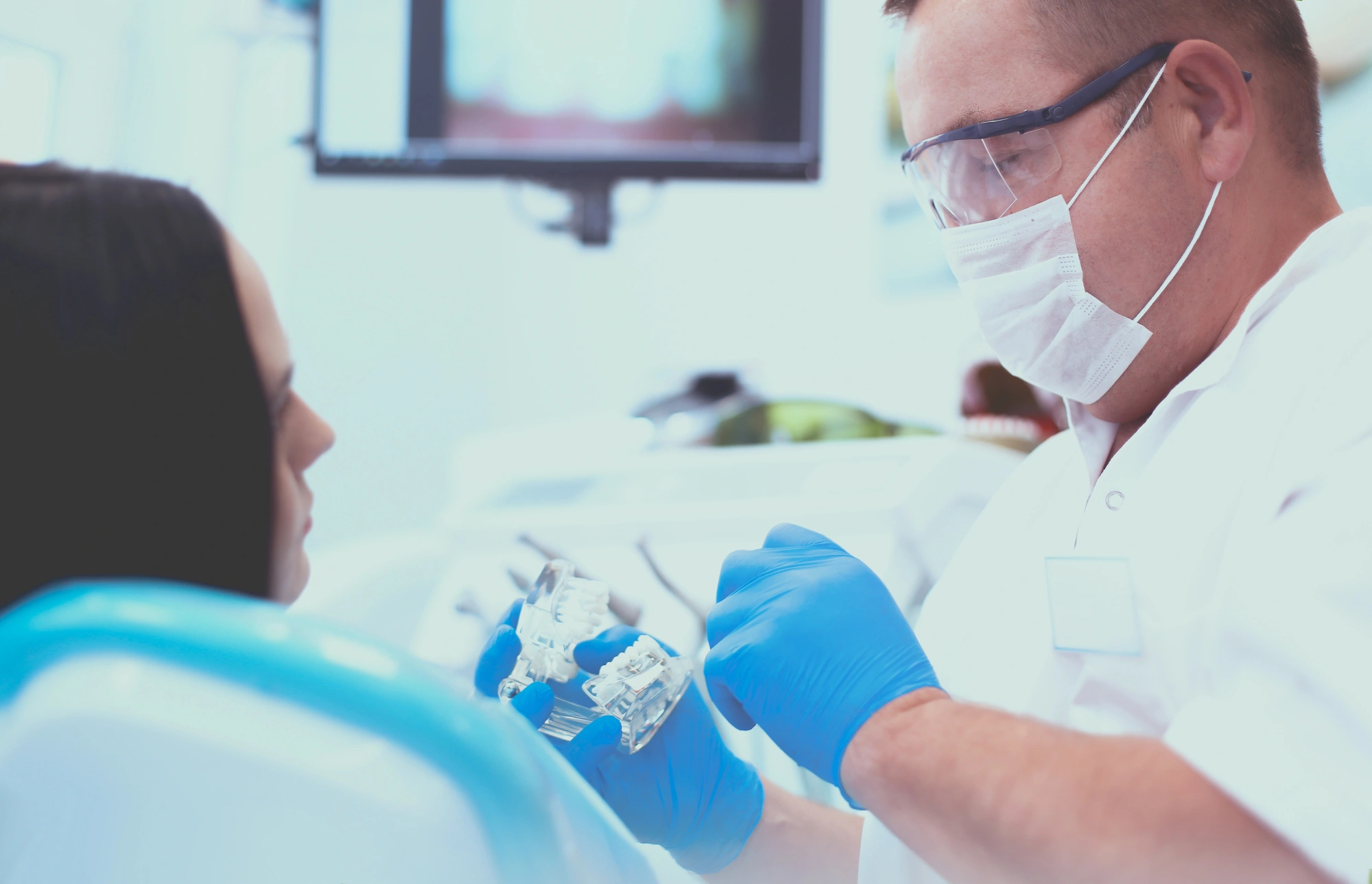 Dentist wearing gloves showing dental models to patient in clinic with dental chair and screen in background