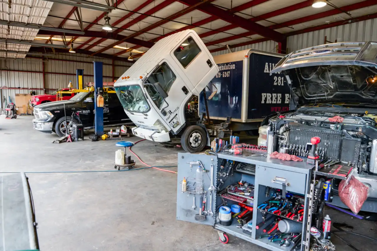 Cab-over heavy-duty truck tilted for engine service; open-hood pickup and trailer reading “FREE” surround tool-laden bay.