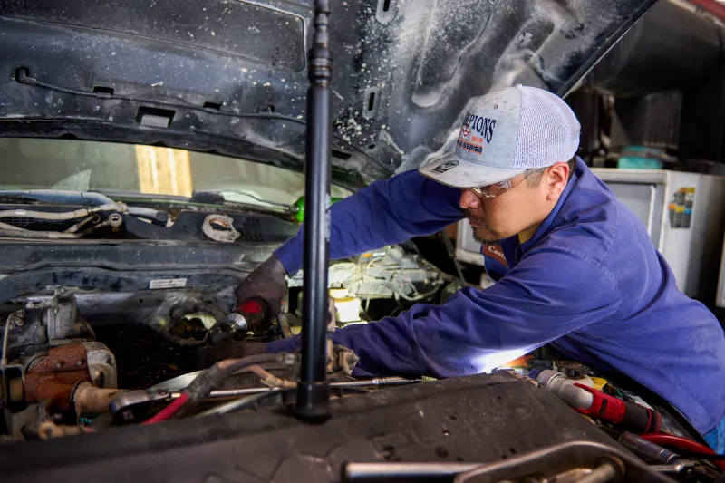 Mechanic in blue uniform working on a truck engine with the hood open inside a repair shop.