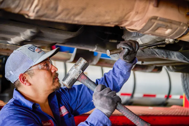 Technician performing exhaust repair under a truck using a hammer and punch tool for pipe alignment.