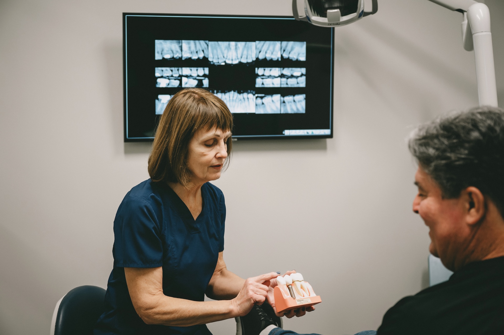Dentist explains dental implant model to patient with x-rays in background