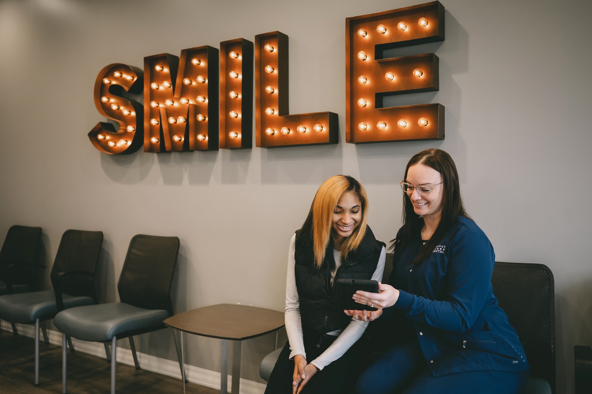 Patients smile while viewing tablet in dental office waiting area under “SMILE” sign