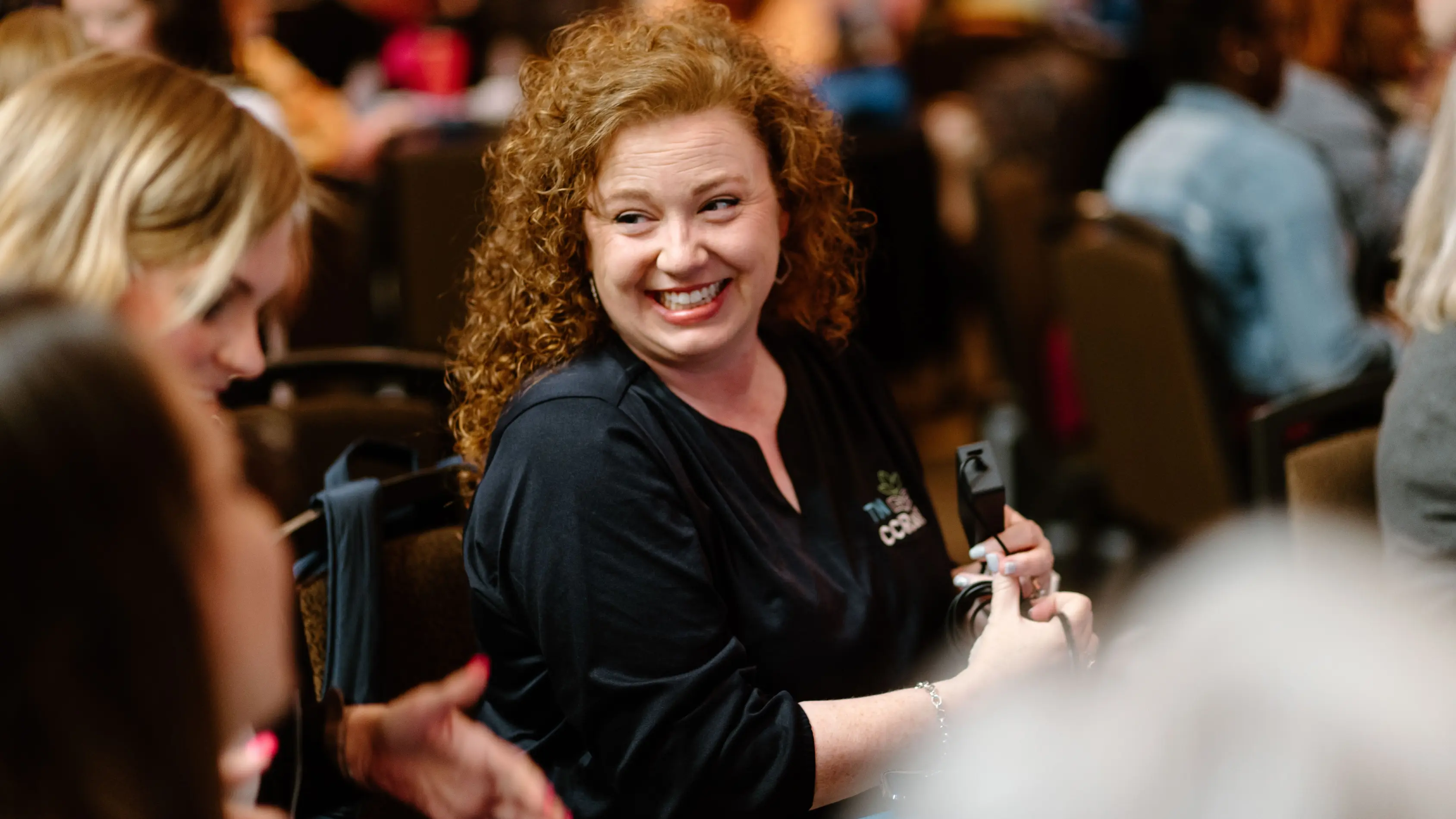 Lady with red hair smiling in a black shirt while holding a black cord.