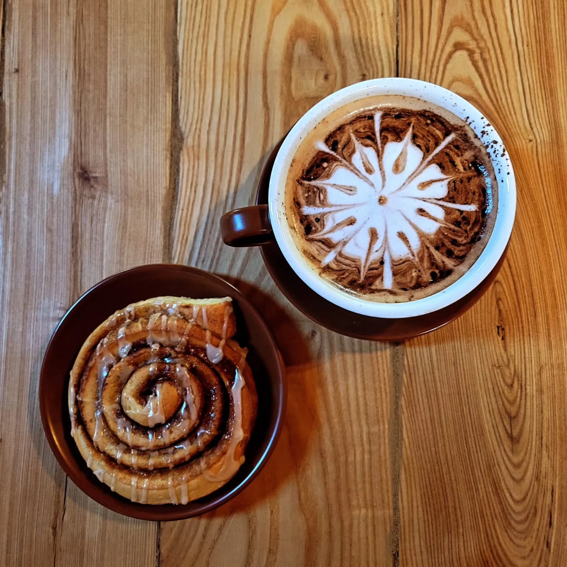 Overhead view of a cinnamon roll with icing on a brown plate next to a cup of coffee with flower-shaped latte art on a wooden table.