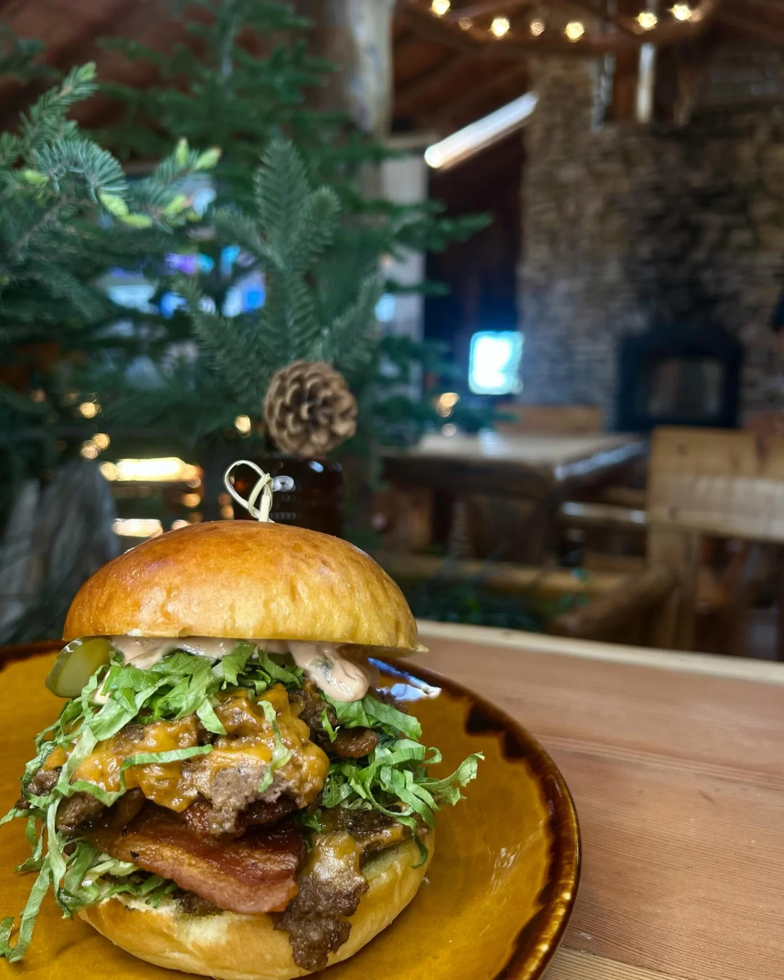 Cheeseburger with bacon, shredded lettuce, pickles, and sauce on a brown plate in a rustic setting.