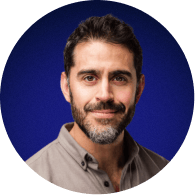 Headshot of a smiling man with dark hair and beard wearing a brown shirt against a blue background.