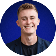 Smiling young man with short blonde hair wearing a black jacket against a blue background.