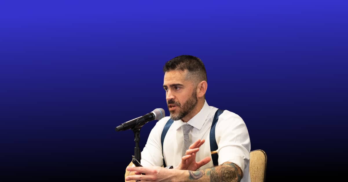Man with tattoos speaking into a microphone against a blue gradient background.