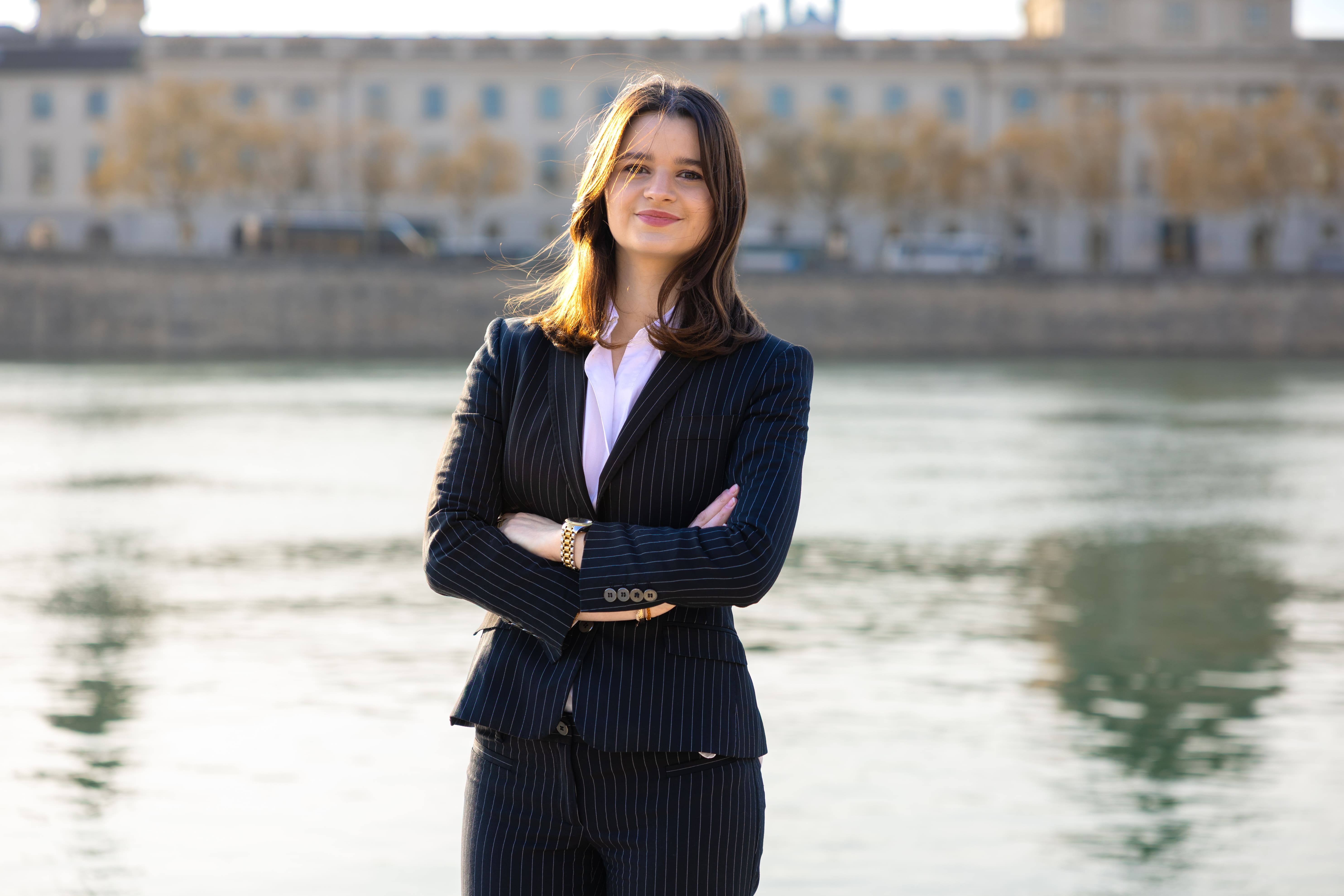Confident young woman in a navy pinstripe suit standing with arms crossed by a riverside with a cityscape background.