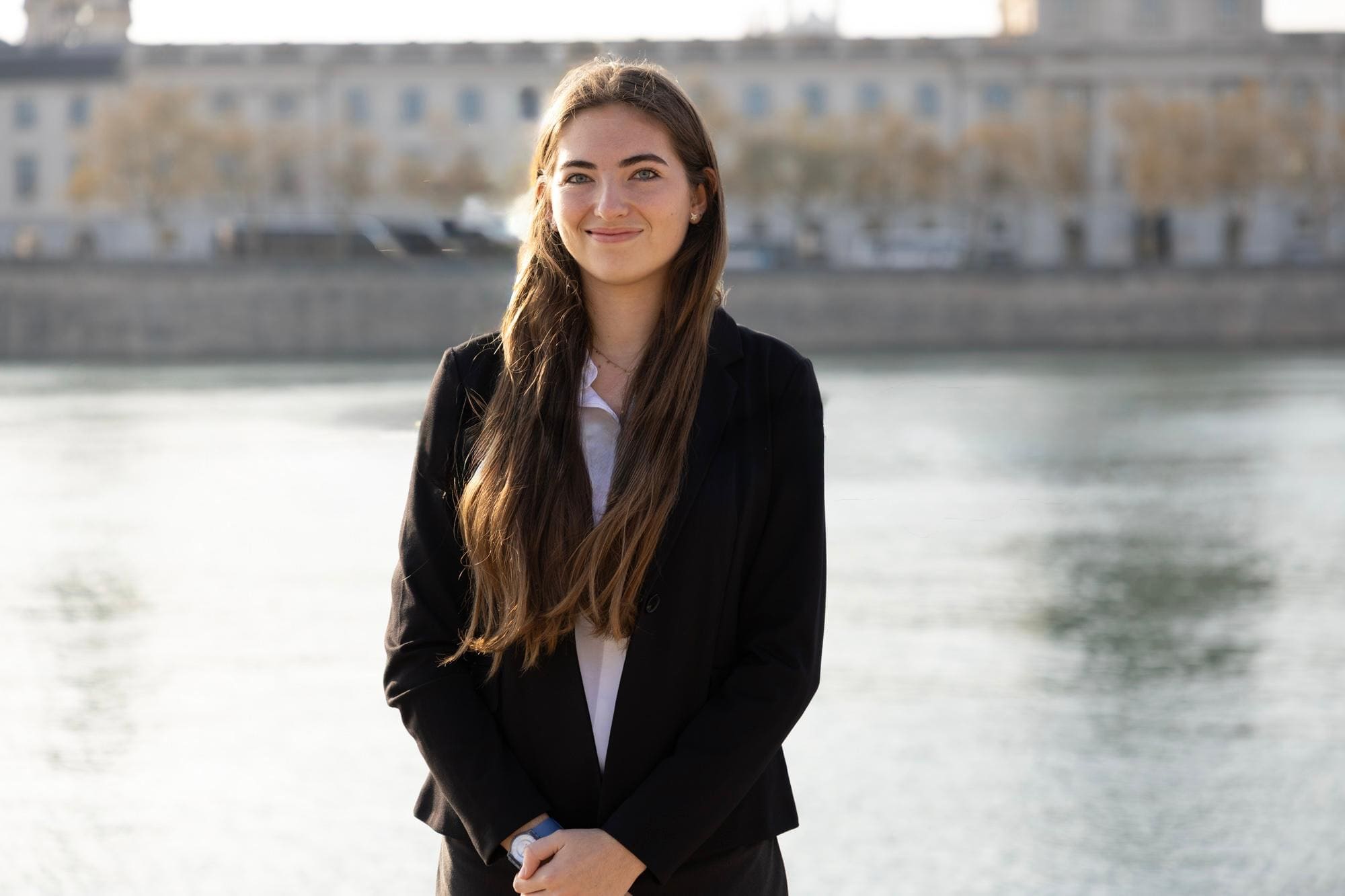 Young woman with long brown hair wearing a black blazer standing by a river with a blurred cityscape background.