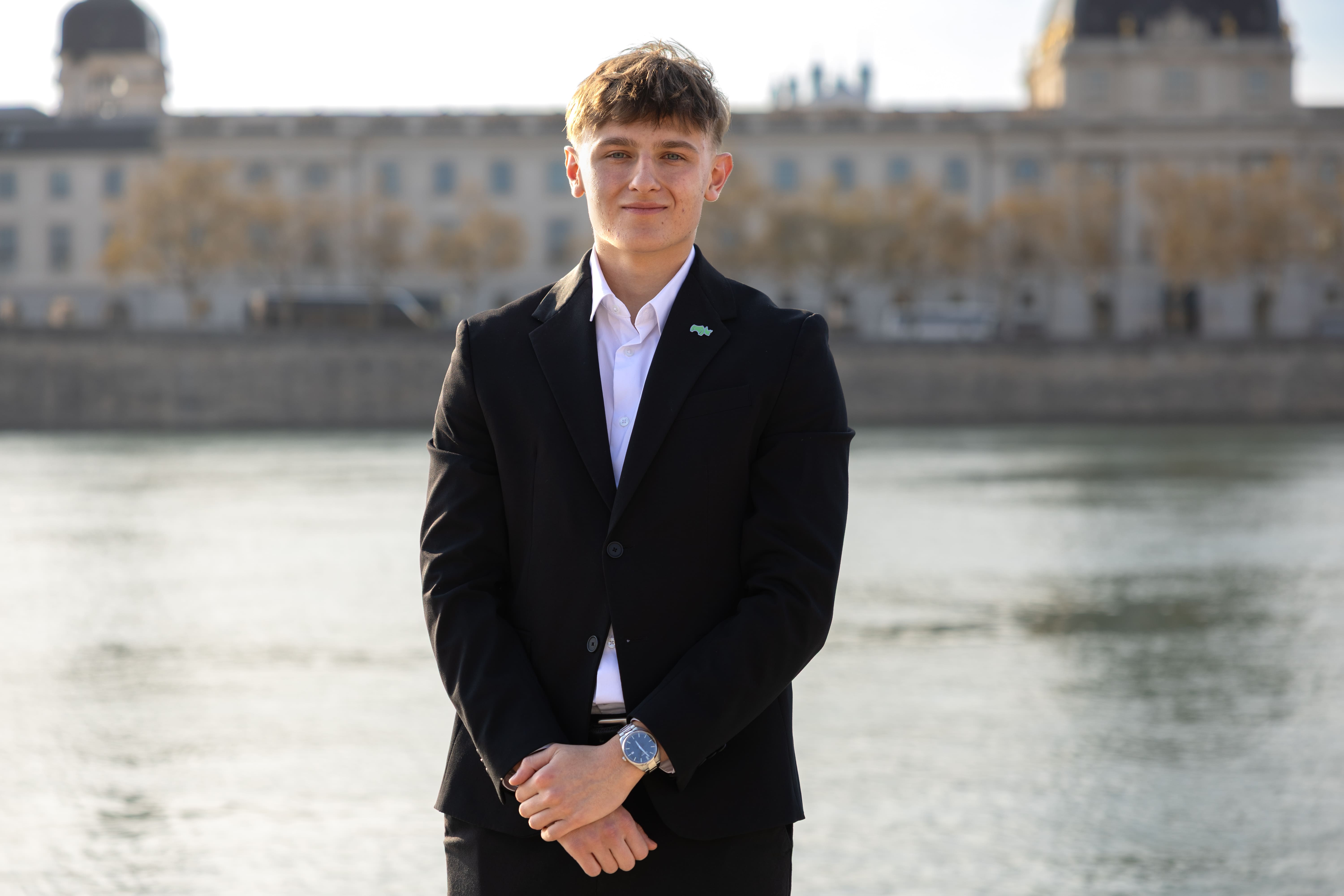 Young man in a black suit and white shirt standing with hands clasped in front of a river and historic building background.