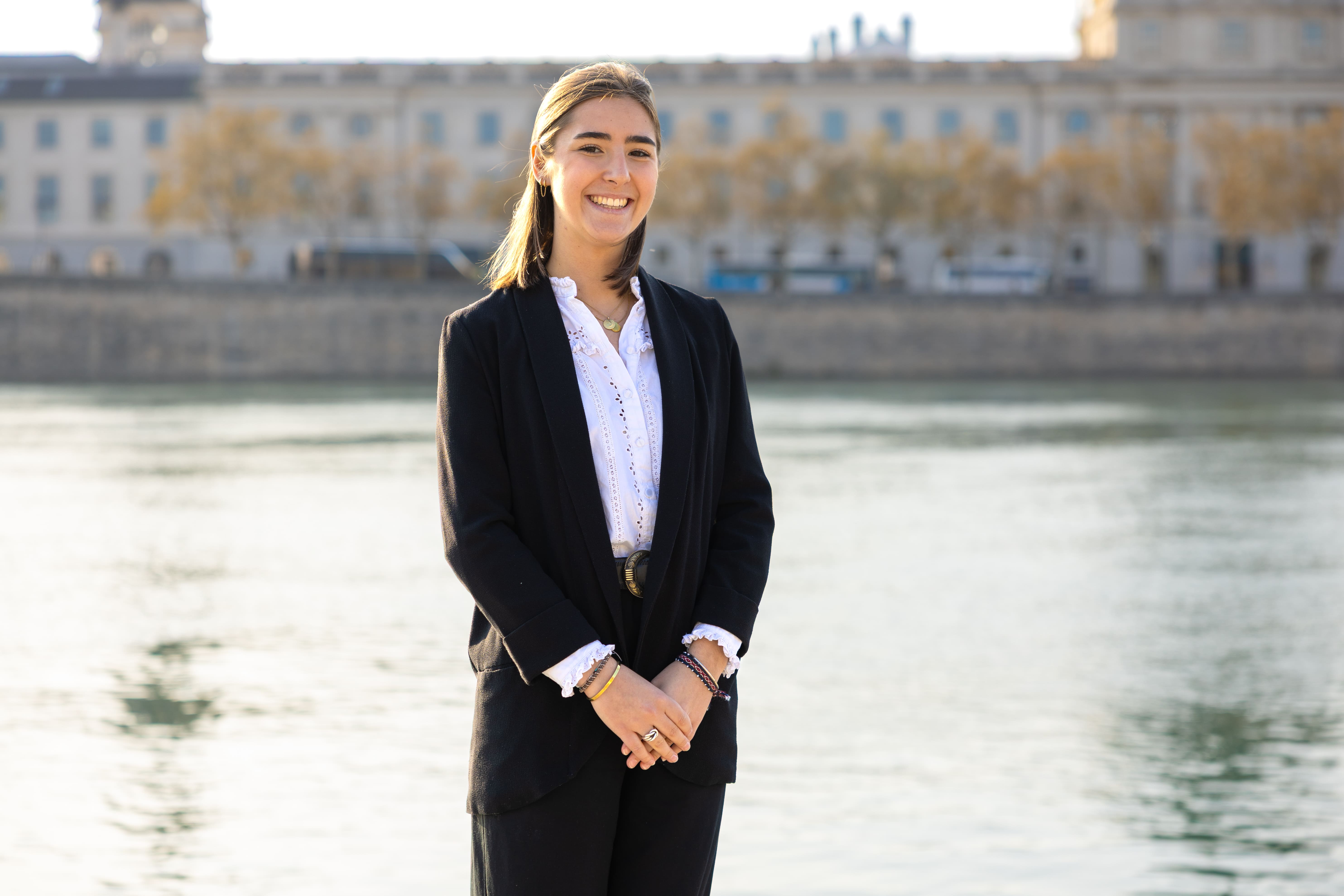 Young woman smiling and standing in front of a river with buildings blurred in the background.