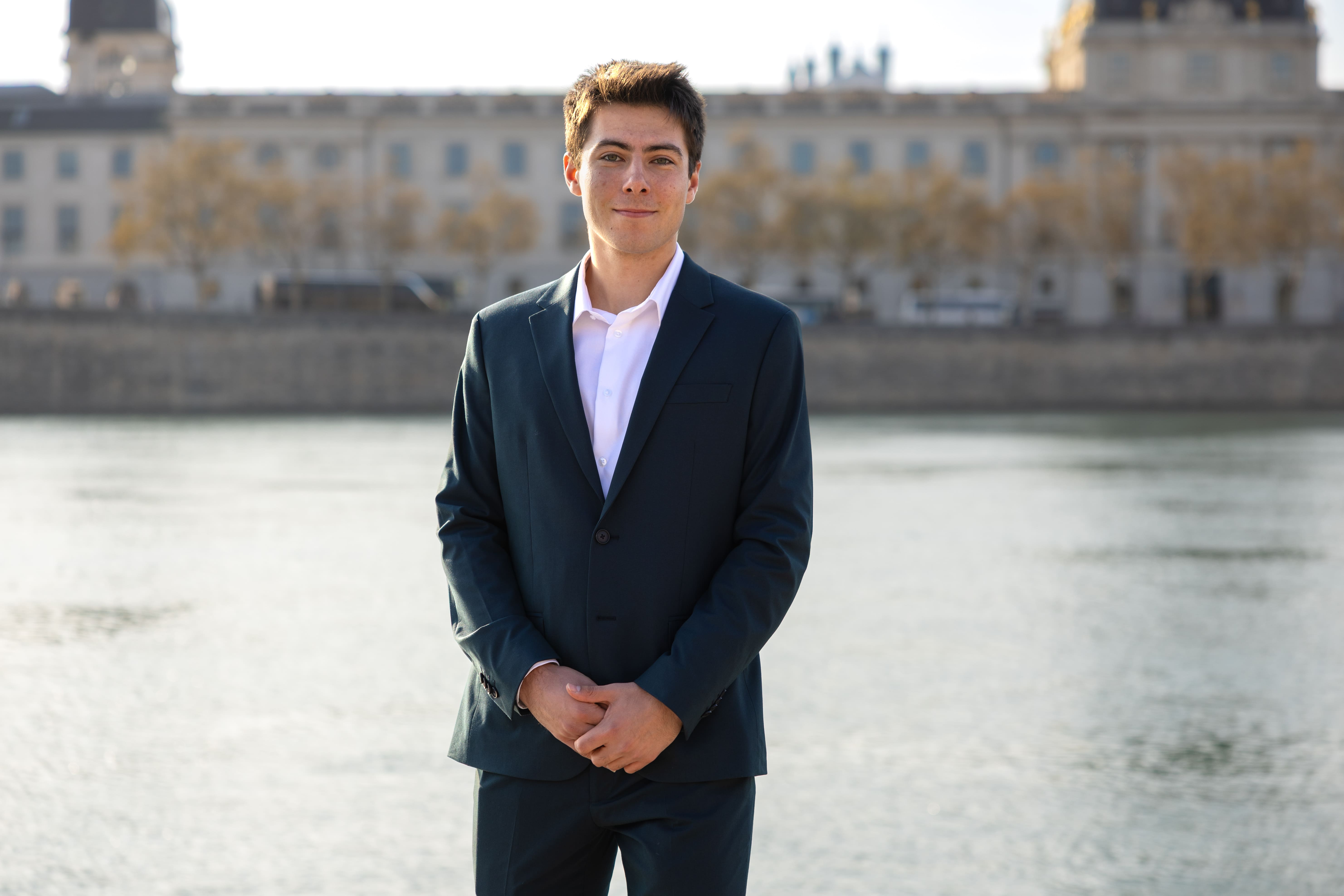 Young man in a dark suit and white shirt standing calmly by a river with a blurred building in the background.
