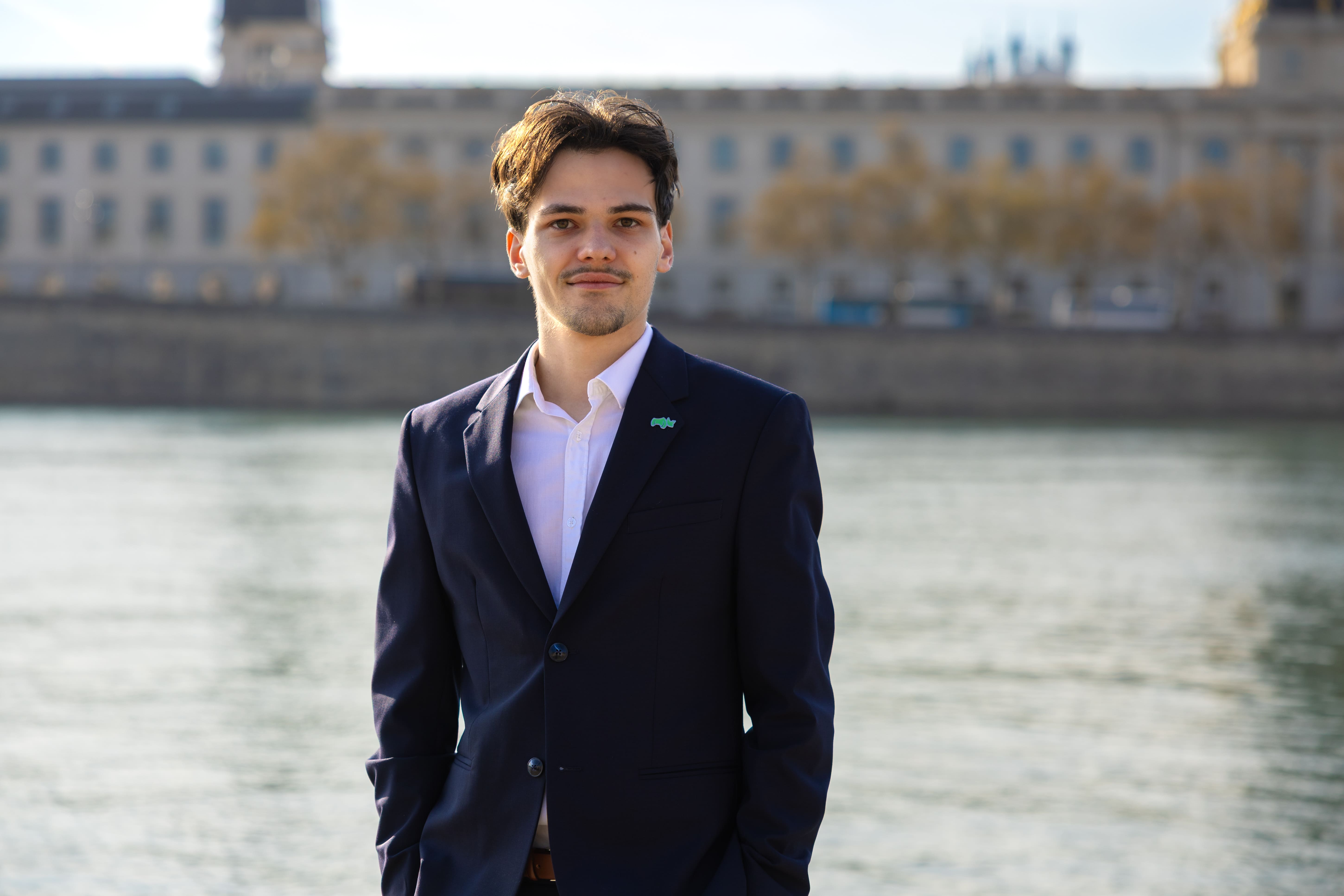 Young man in a navy blazer and white shirt standing by a waterfront with blurred buildings in the background.