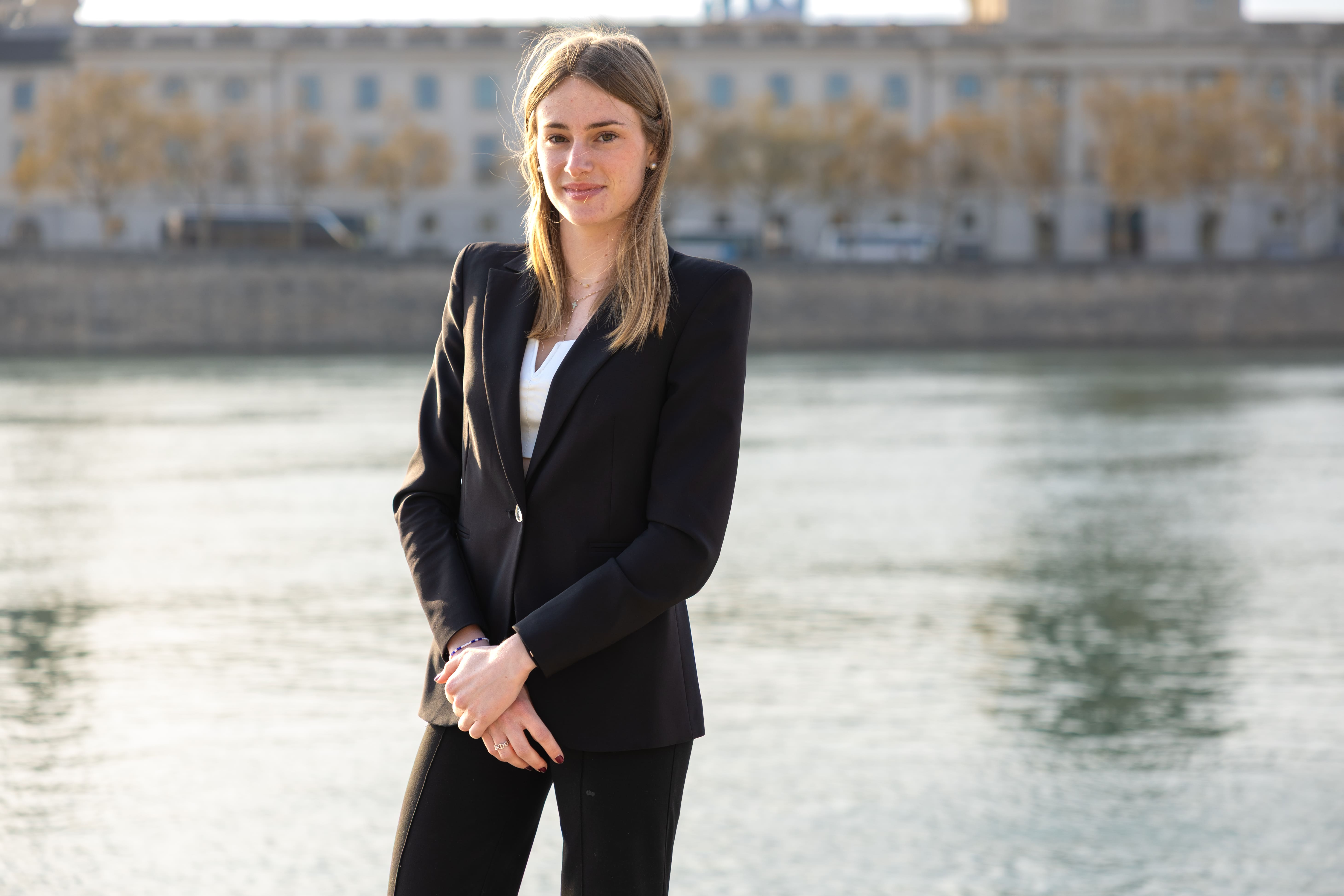 Young woman with blonde hair wearing a black blazer and white top standing by a river with a cityscape in the background.